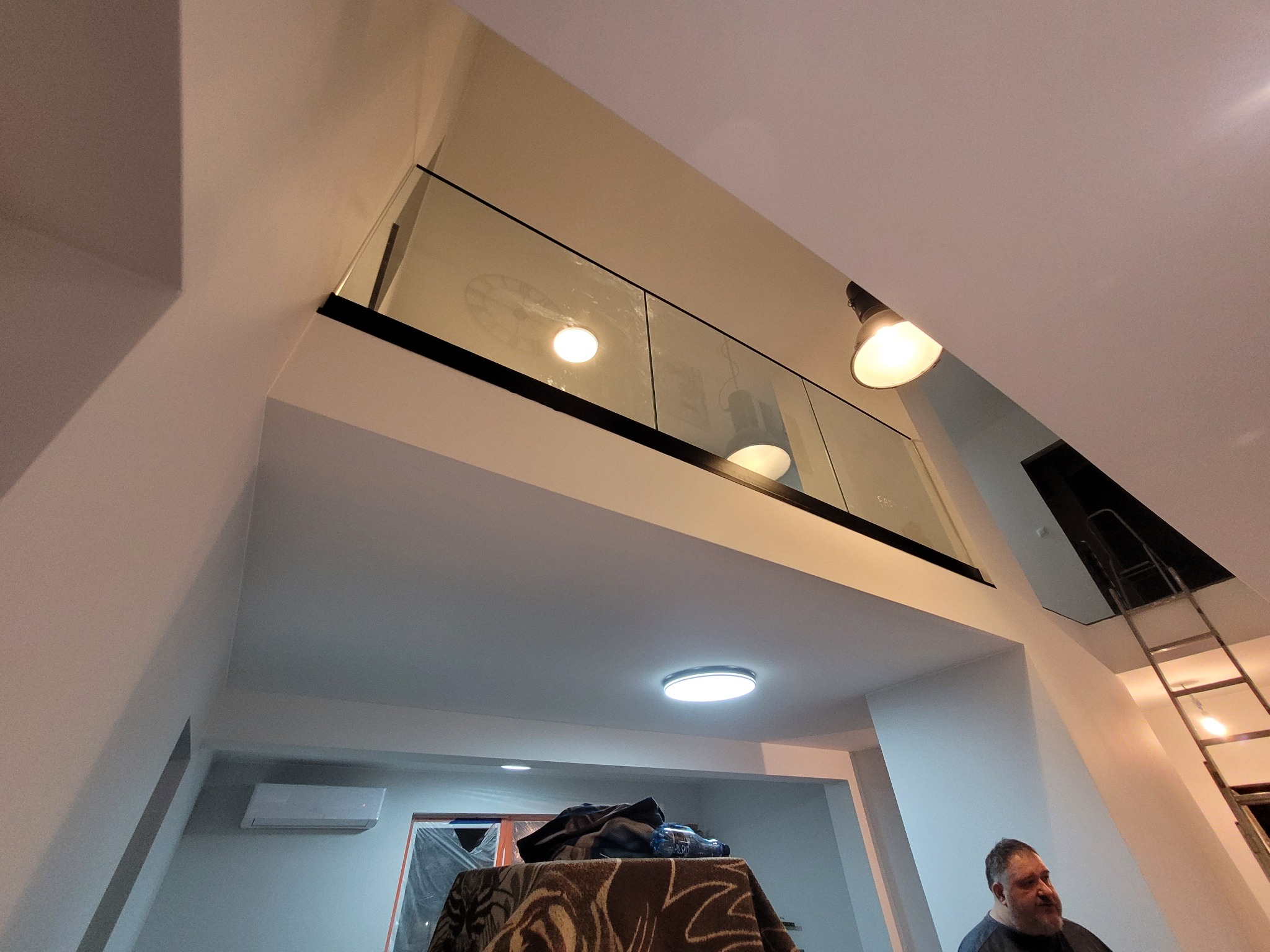 Szklana balustrada z czarnymi profilami w dwupoziomowym wnętrzu w Krzeszowicach, widok z dołu na antresolę, widoczne lampy i drabina.