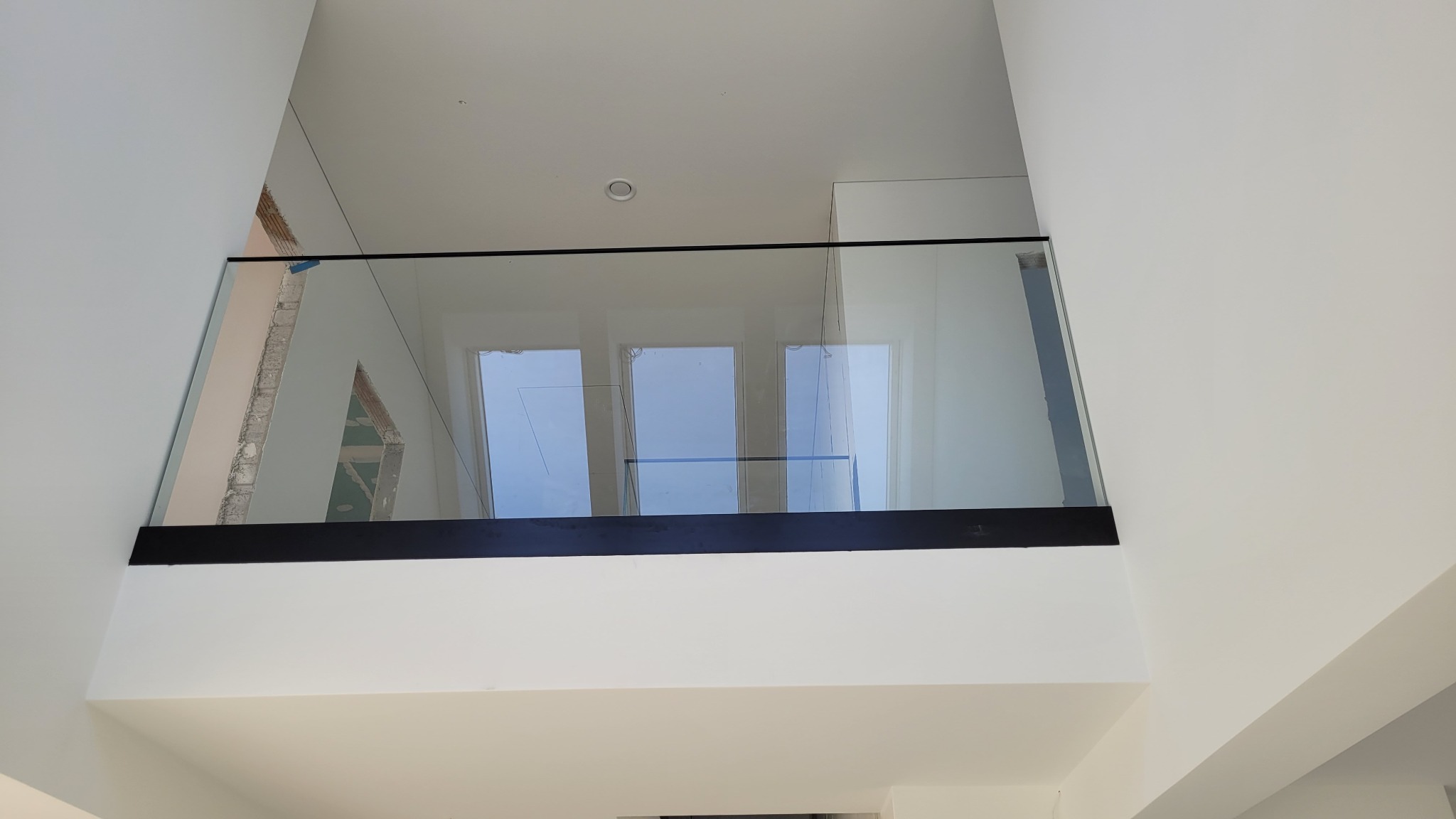 Wewnętrzna balustrada ze szkła z czarną ramą, widok z dołu na górę, widoczne okna w tle i surowy otwór w ścianie.