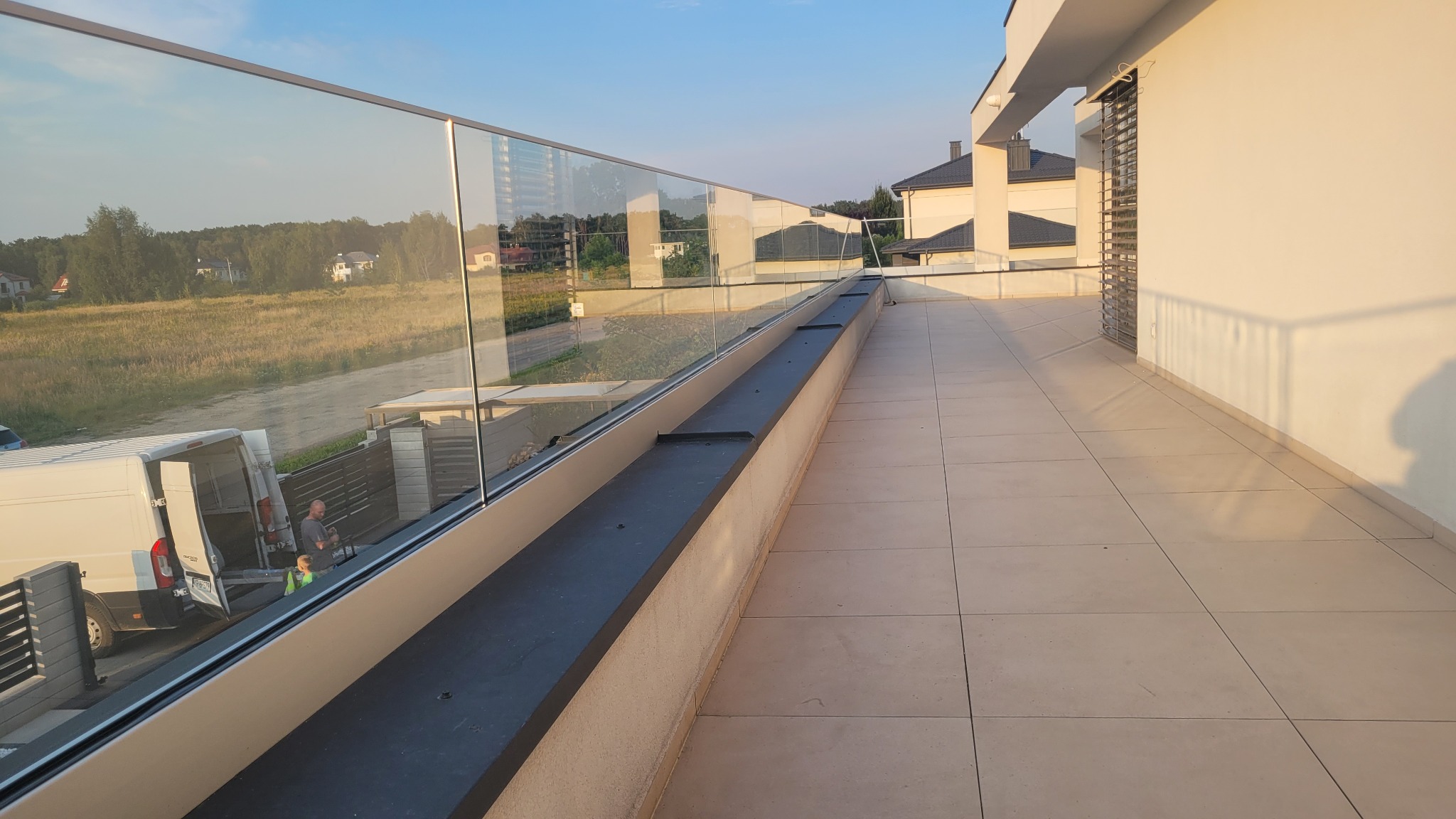 Nowoczesny balkon z szklaną balustradą, widok na okolicę z odbijającym się w szkle krajobrazem i pracownikami przy furgonetce w tle.
