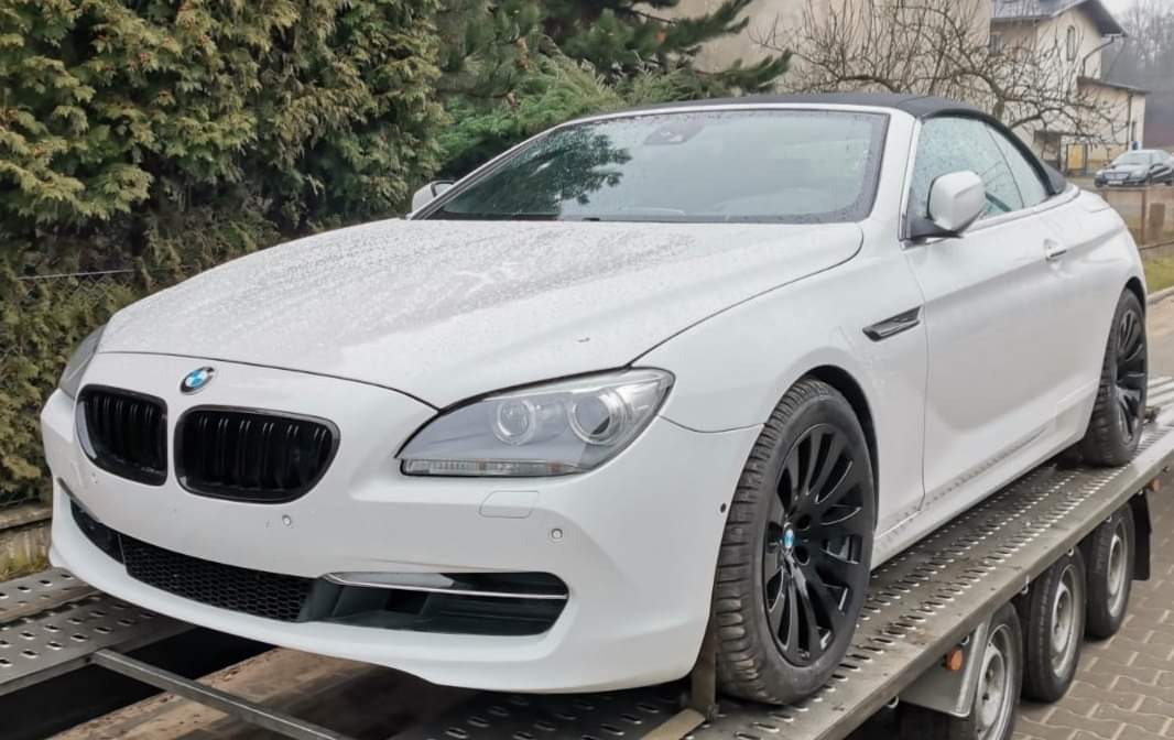 Białe BMW kabriolet z czarnymi felgami transportowane na niskopodwoziowej lawecie, widoczne krople deszczu na karoserii.