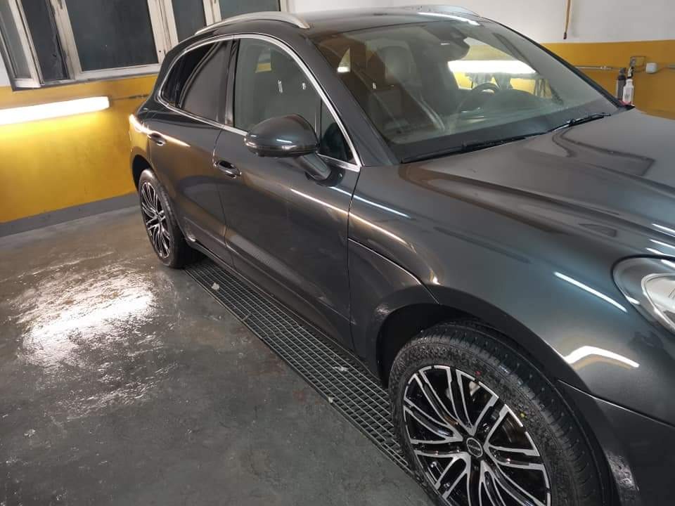 Szary SUV marki Porsche Macan po umyciu, z widocznymi kroplami wody na lakierze, stojący w pomieszczeniu z żółtą ścianą i metalową kratką pod kołami.