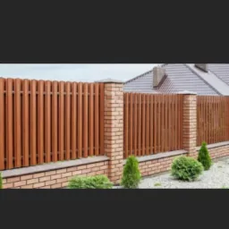 Brązowe, pionowe panele ogrodzeniowe z ceglanymi słupkami i podmurówką, otoczone zielonymi krzewami i małymi drzewkami iglastymi, na tle domu z szarą dachówką.