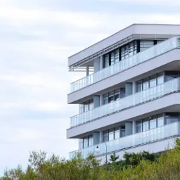 Nowoczesny, biały apartamentowiec z przeszklonymi balkonami, otoczony zielenią, widziany z dołu pod lekkim kątem.