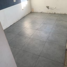 MICHAŁ DEMBEK USŁUGI BUDOWLANE - Szare, kwadratowe płytki podłogowe ułożone w pomieszczeniu w trakcie remontu, widoczne ślady po pracach na ścianach i instalacja hydrauliczna.