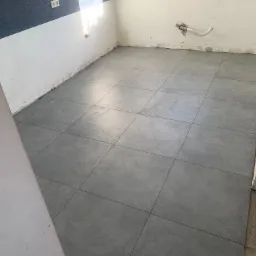 Szare, kwadratowe płytki podłogowe ułożone w pomieszczeniu w trakcie remontu, widoczne ślady po pracach na ścianach i instalacja hydrauliczna.