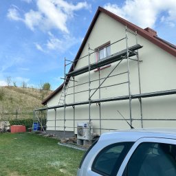 MICHAŁ DEMBEK USŁUGI BUDOWLANE - Dom jednorodzinny z jasną elewacją, na tle błękitnego nieba z chmurami. Widoczne rusztowania i fragment samochodu. Świeżo otynkowana fasada.