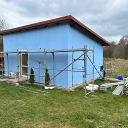 MICHAŁ DEMBEK USŁUGI BUDOWLANE - Niebieski dom z brązowym dachem w trakcie tynkowania, otoczony rusztowaniami i zieloną trawą. Widoczne narzędzia i materiały budowlane. Pochmurne niebo w tle.