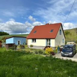 Jasny dom z czerwoną dachówką i oknami dachowymi, otoczony zielenią. Na podjeździe zaparkowane dwa samochody, w tle błękitny budynek i pagórkowaty krajobraz.