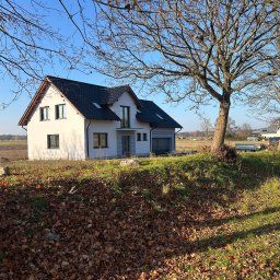 MICHAŁ DEMBEK USŁUGI BUDOWLANE - Nowoczesny dom jednorodzinny z garażem, wykończony w bieli, z ciemną dachówką, na tle jesiennego krajobrazu. Widoczne drzewa bez liści i opadłe liście.