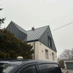 MICHAŁ DEMBEK USŁUGI BUDOWLANE - Nowoczesny dom z szarą blachą trapezową na dachu, kremową elewacją i minimalistycznymi lampami zewnętrznymi, częściowo zasłonięty przez drzewa i samochód.