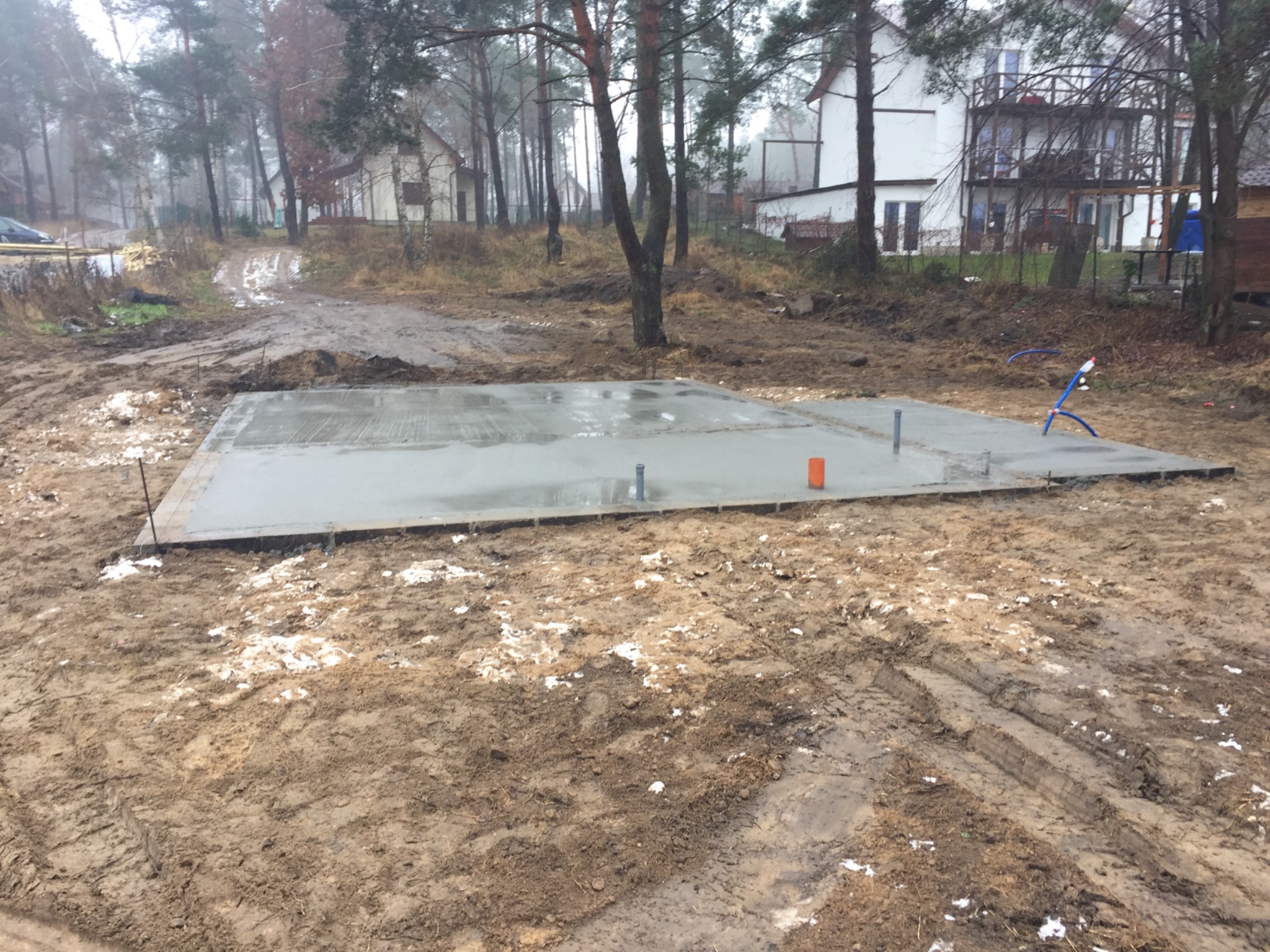 Świeżo wylana, mokra płyta fundamentowa pod budynek w surowym otoczeniu, widoczne wystające rury i pomarańczowy słupek geodezyjny, w tle domy i drzewa.