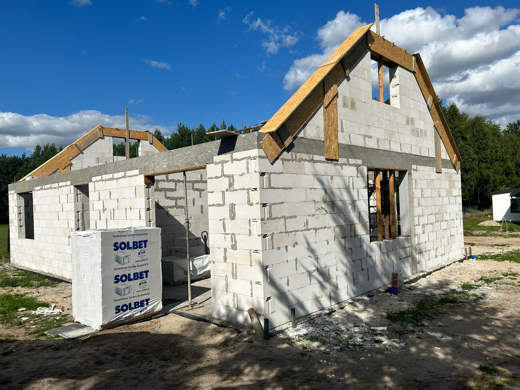 Budowa domu w stanie surowym otwartym z bloczków Solbet, widoczne elementy więźby dachowej i paleta bloczków na placu budowy.