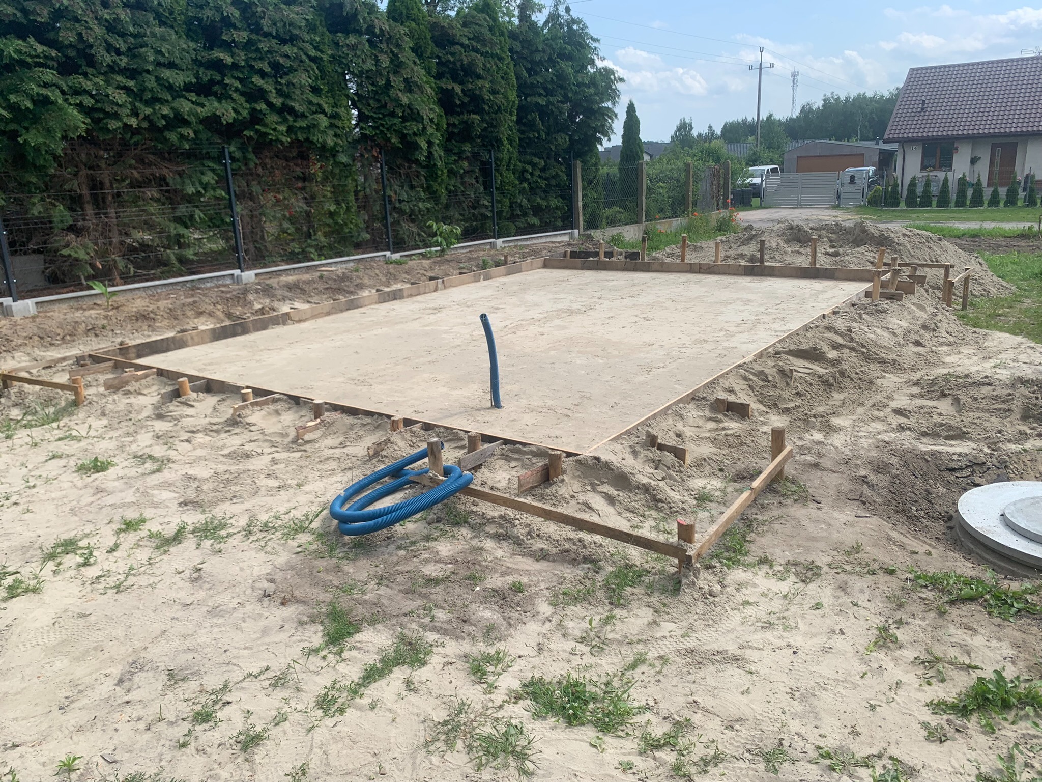 Przygotowany teren pod budowę: wyrównany piasek otoczony drewnianym szalunkiem, widoczne przyłącze wodne w postaci niebieskiego węża, w tle ogrodzenie i zieleń.