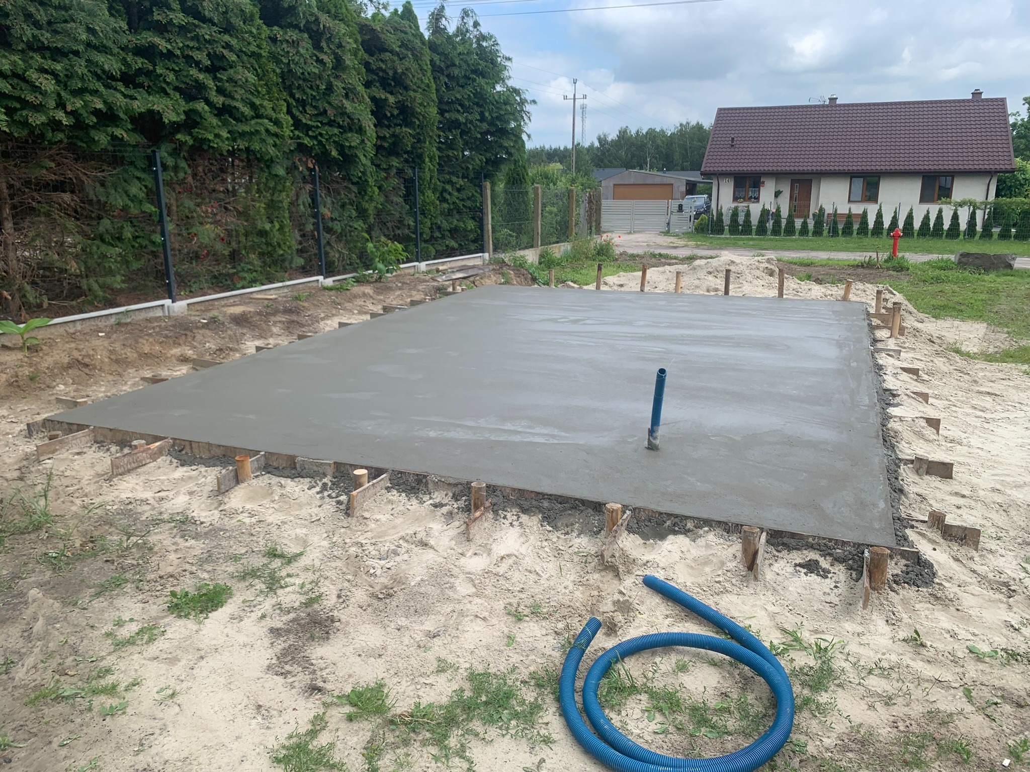Wylany beton na fundament domu jednorodzinnego z widocznymi drewnianymi szalunkami i niebieskim wężem do wody leżącym obok.