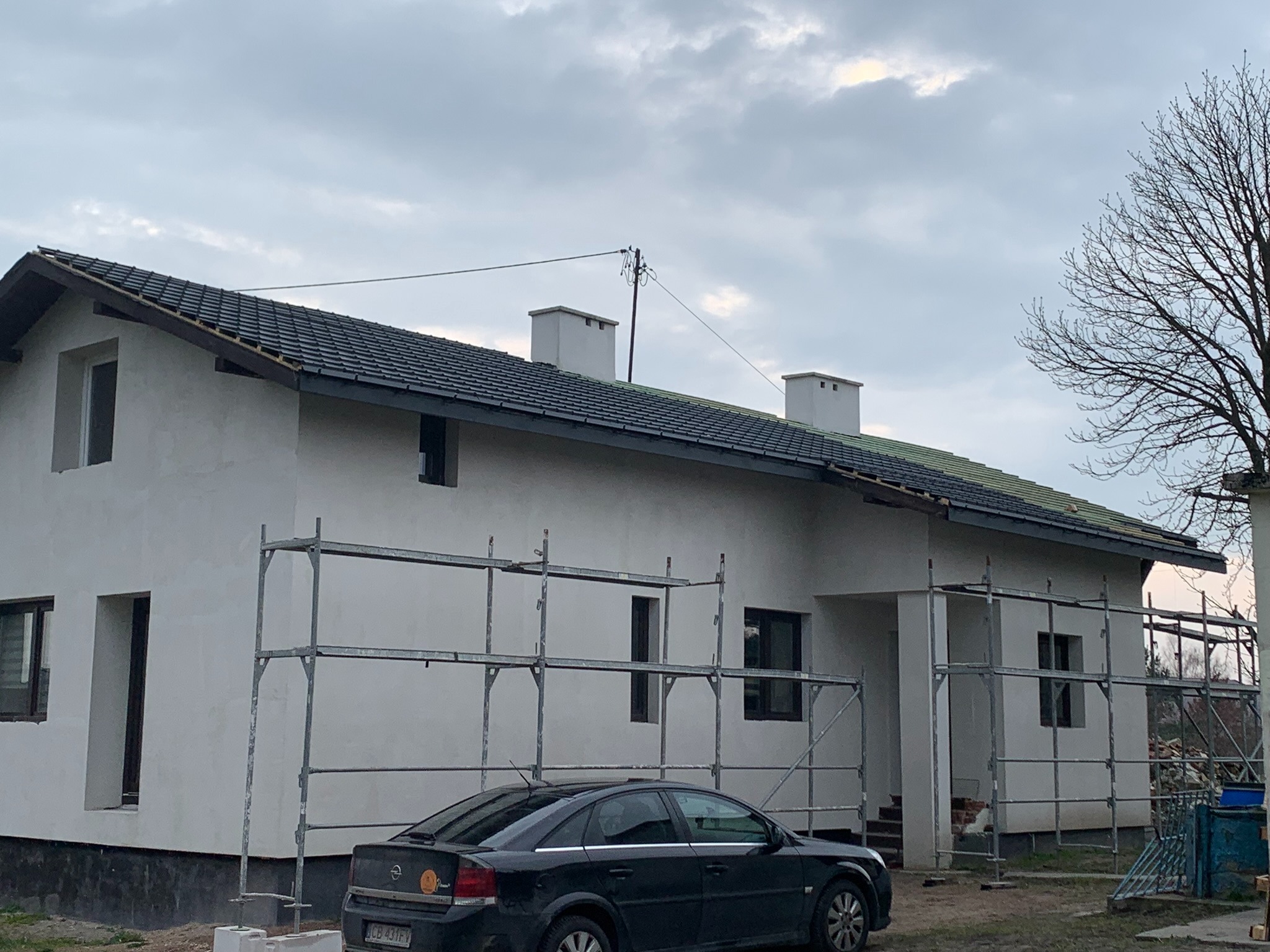 Dom jednorodzinny w trakcie prac wykończeniowych elewacji, z widocznymi rusztowaniami i częściowo położonym dachem z ciemnej dachówki. Elewacja otynkowana na biało, okna w ciemnych ramach.