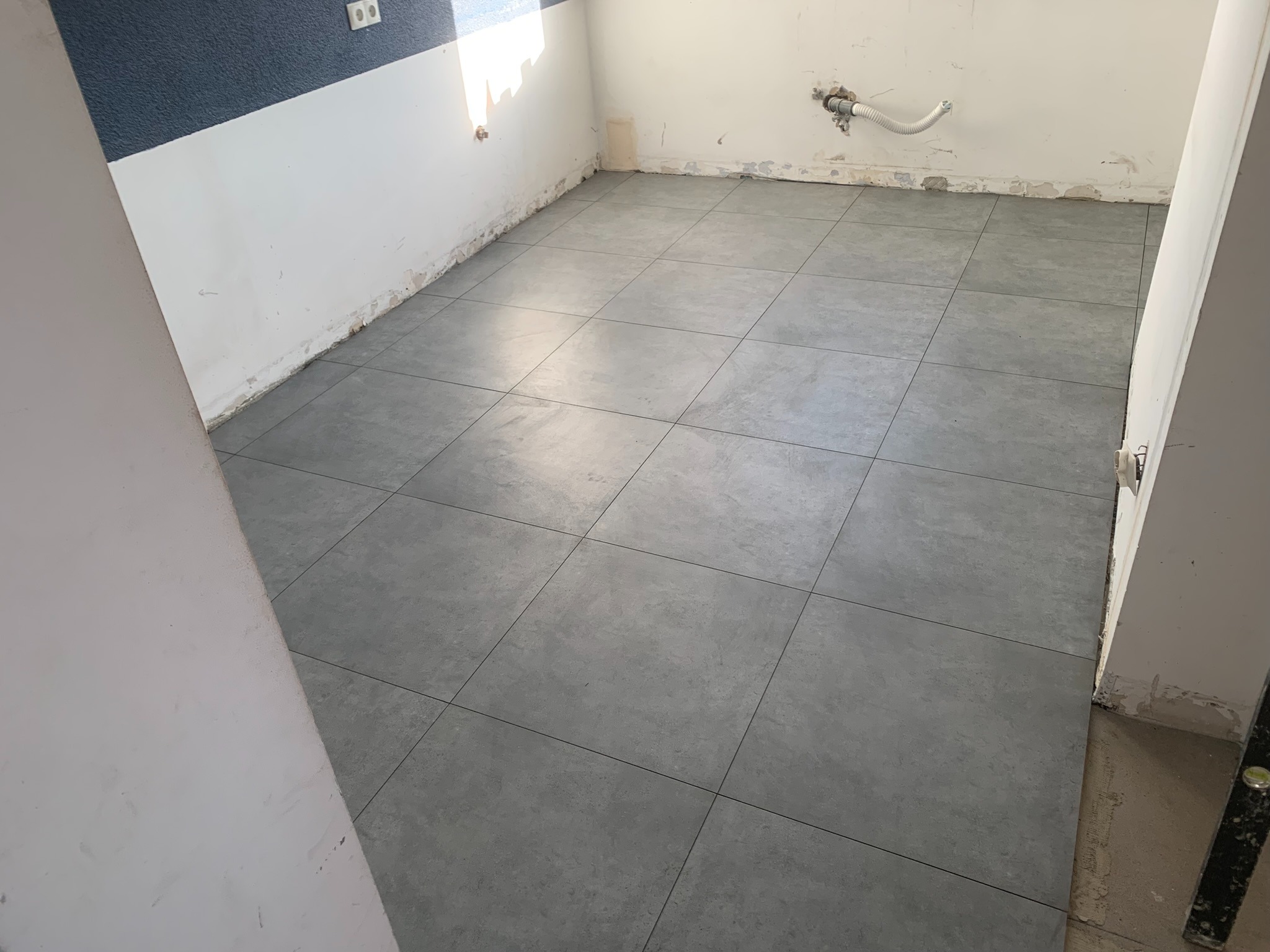 Szare, kwadratowe płytki podłogowe ułożone w pomieszczeniu w trakcie remontu, widoczne ślady po pracach na ścianach i instalacja hydrauliczna.