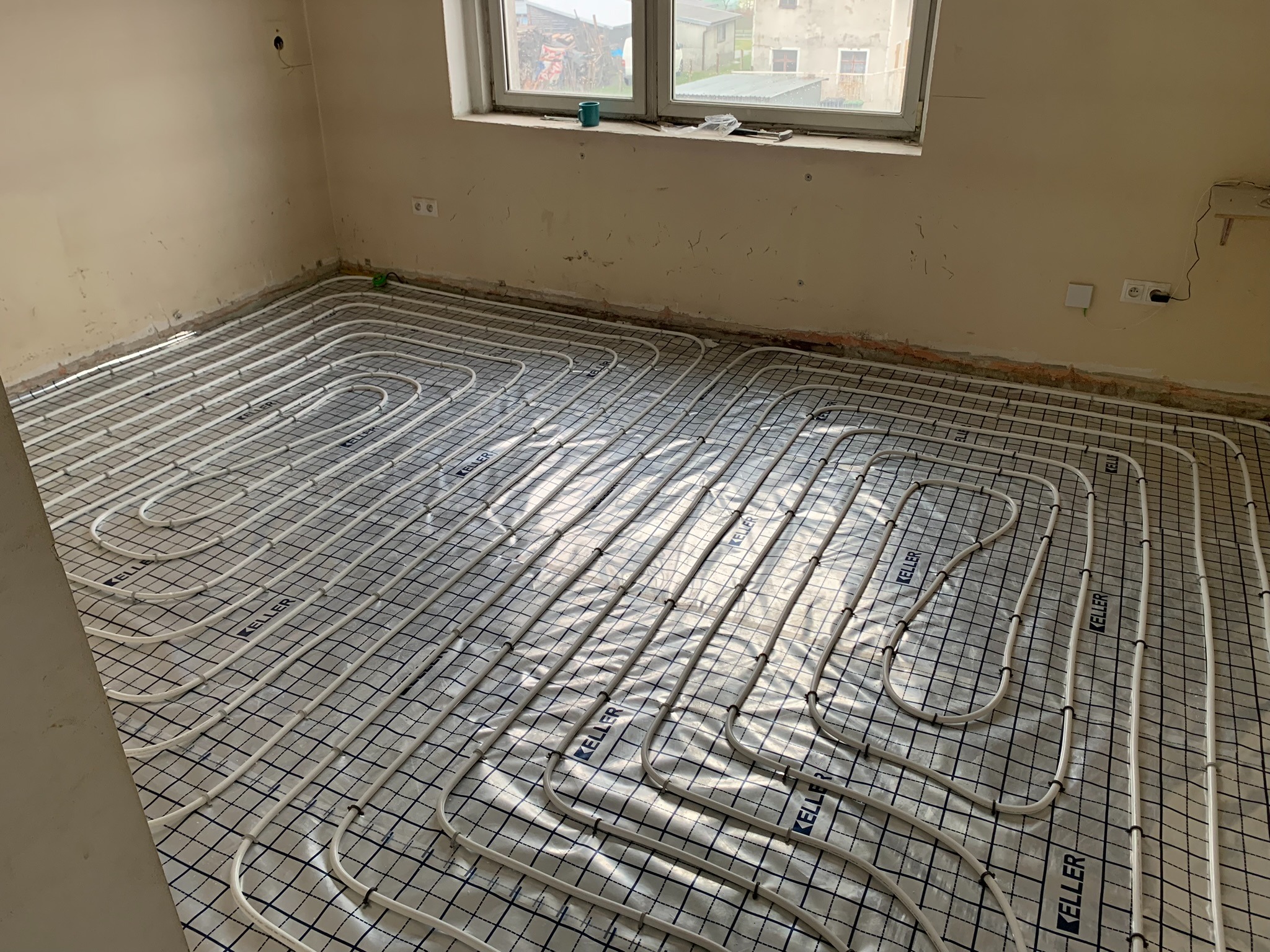 Białe rurki ogrzewania podłogowego marki Keller, ułożone na folii z nadrukowaną siatką, w surowym pomieszczeniu z oknem.