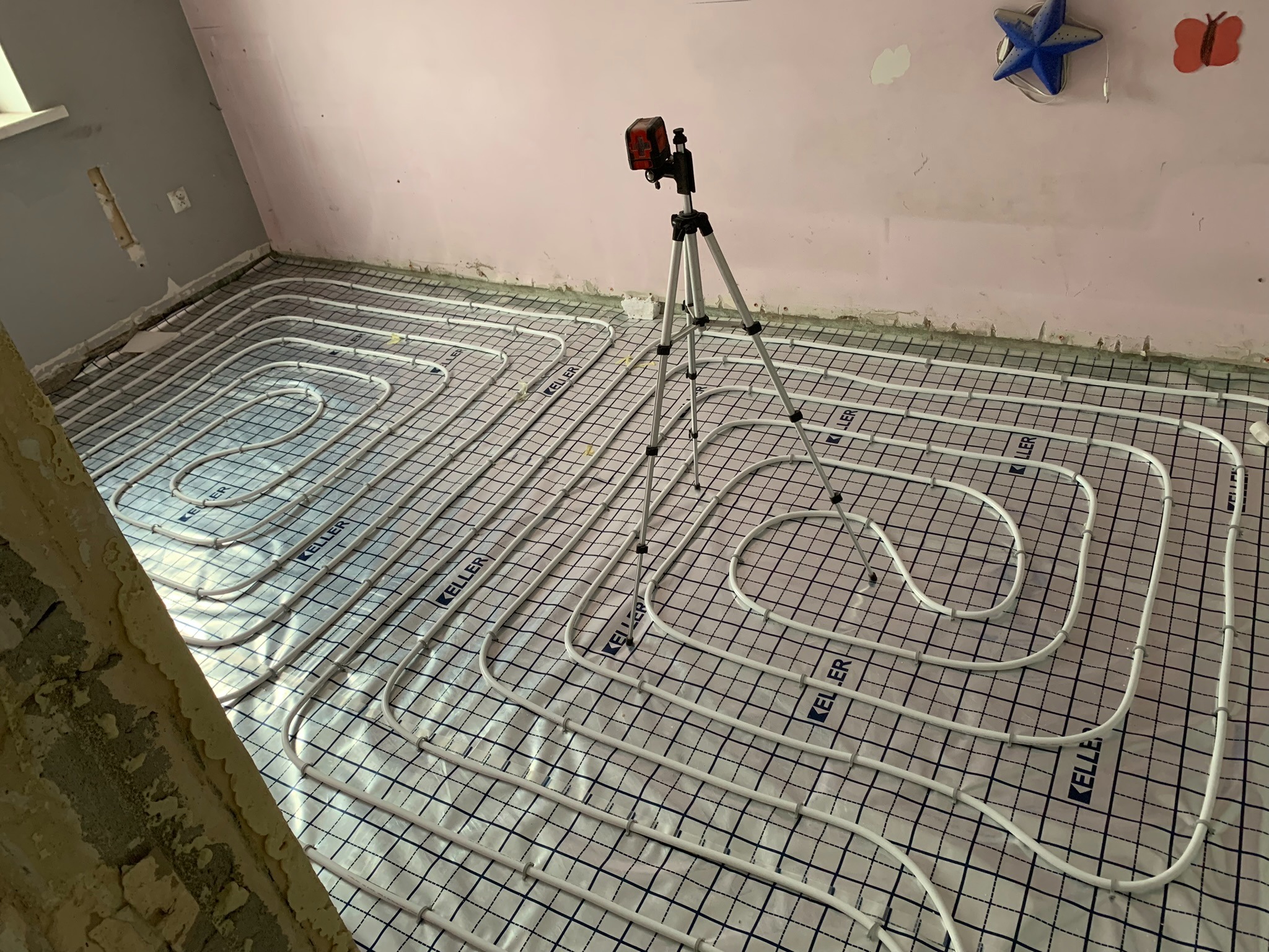 Rozłożone białe rury ogrzewania podłogowego na folii z nadrukiem kratki, poziomowanie laserowe na statywie w pokoju z różowymi ścianami w trakcie remontu.