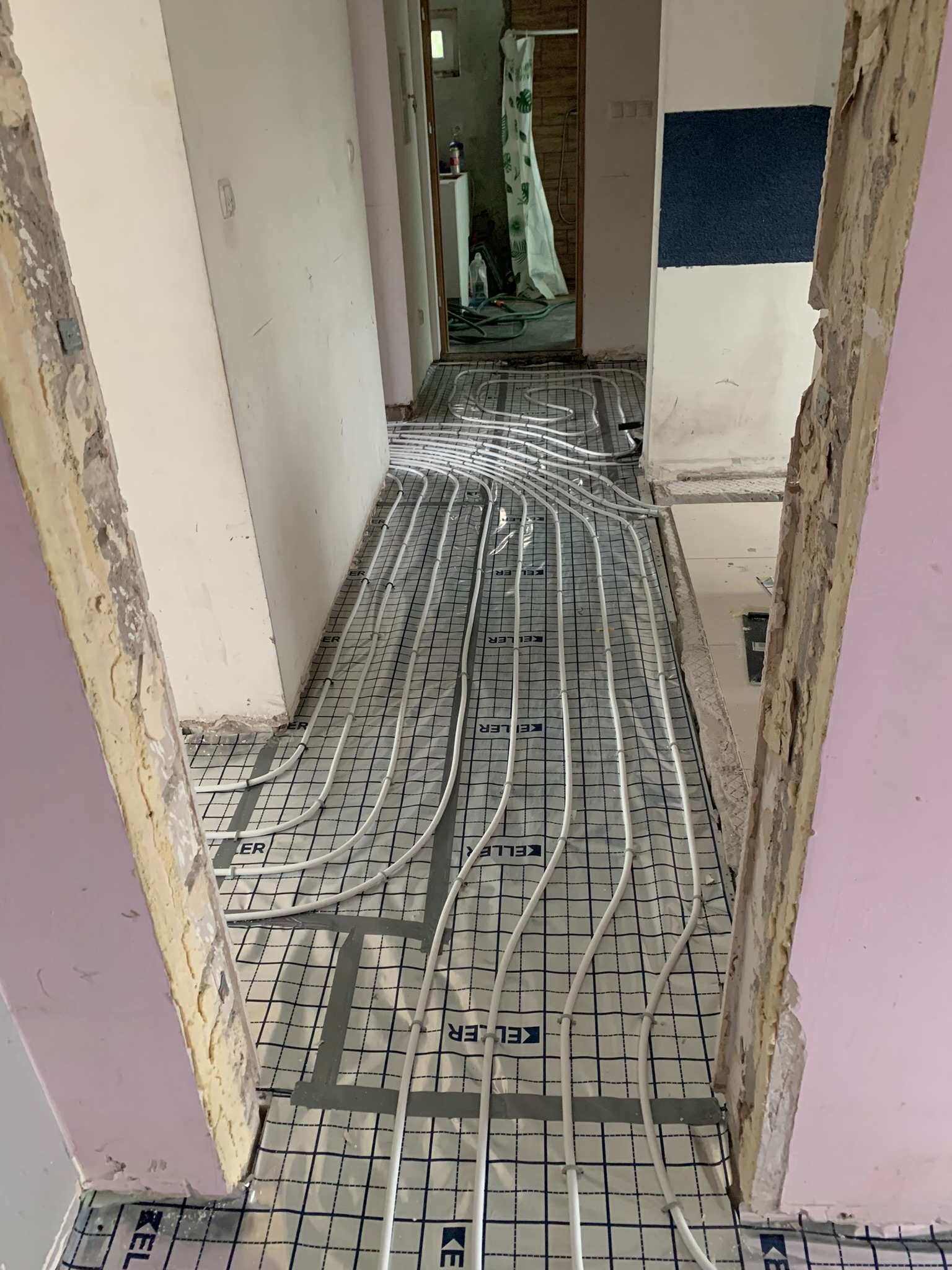 Montaż ogrzewania podłogowego: białe rurki na czarnej siatce z logo Keller, widoczne surowe ściany i futryny przed wykończeniem.