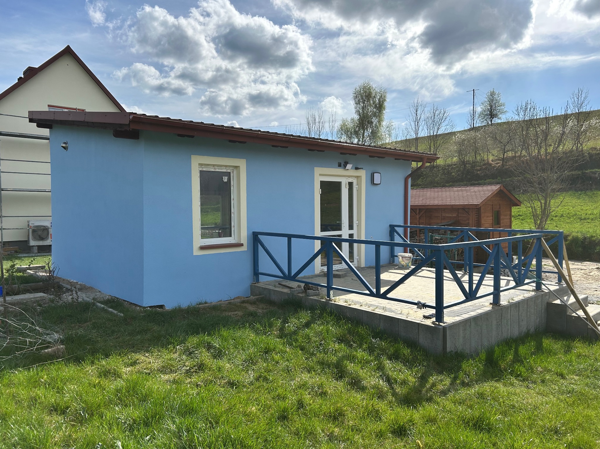 Niebieski, parterowy budynek z tarasem na betonowej podmurówce. Widoczne okno z kremową ramą i białe drzwi. W tle zielony krajobraz.