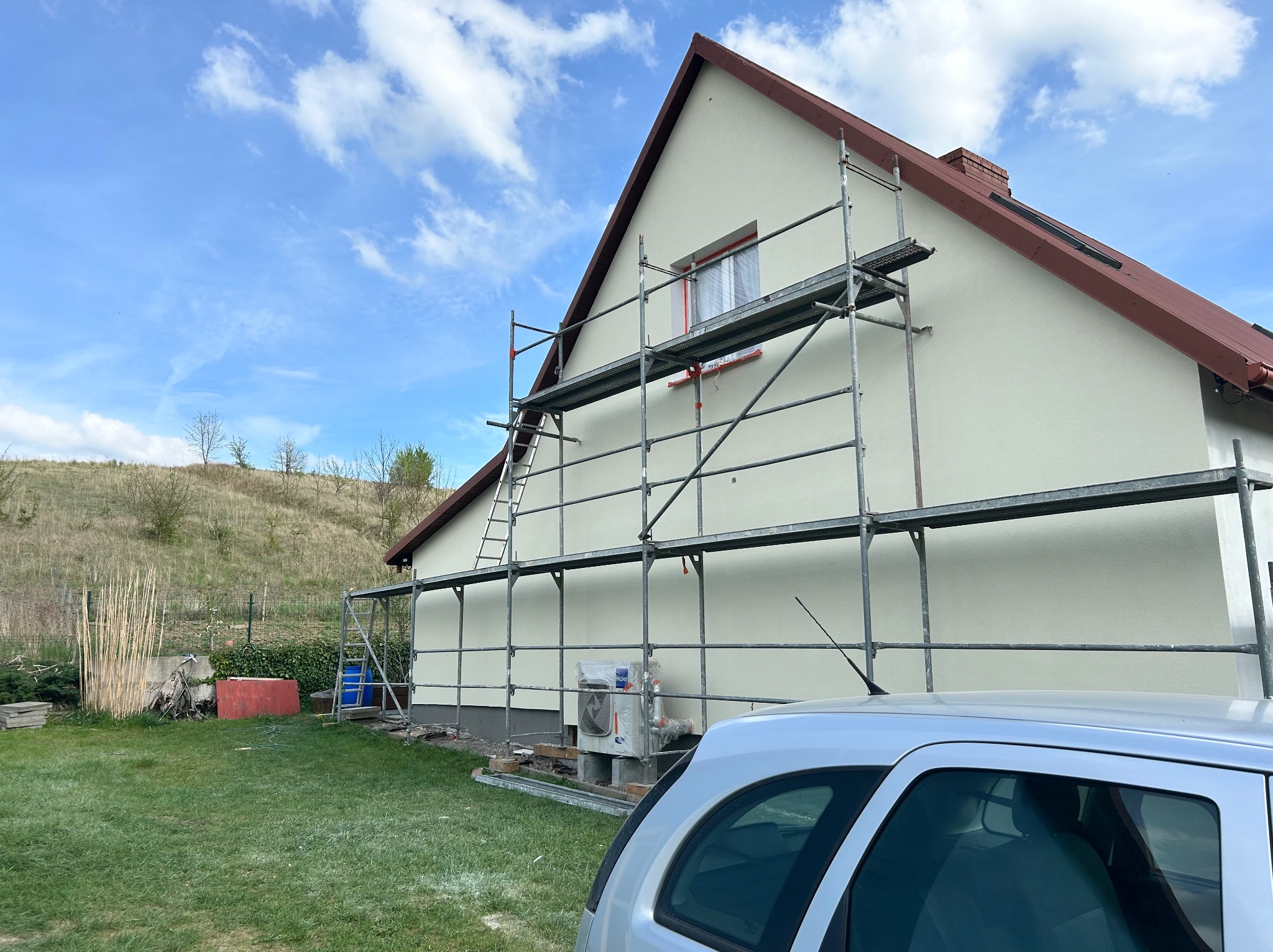 Dom jednorodzinny z jasną elewacją, na tle błękitnego nieba z chmurami. Widoczne rusztowania i fragment samochodu. Świeżo otynkowana fasada.