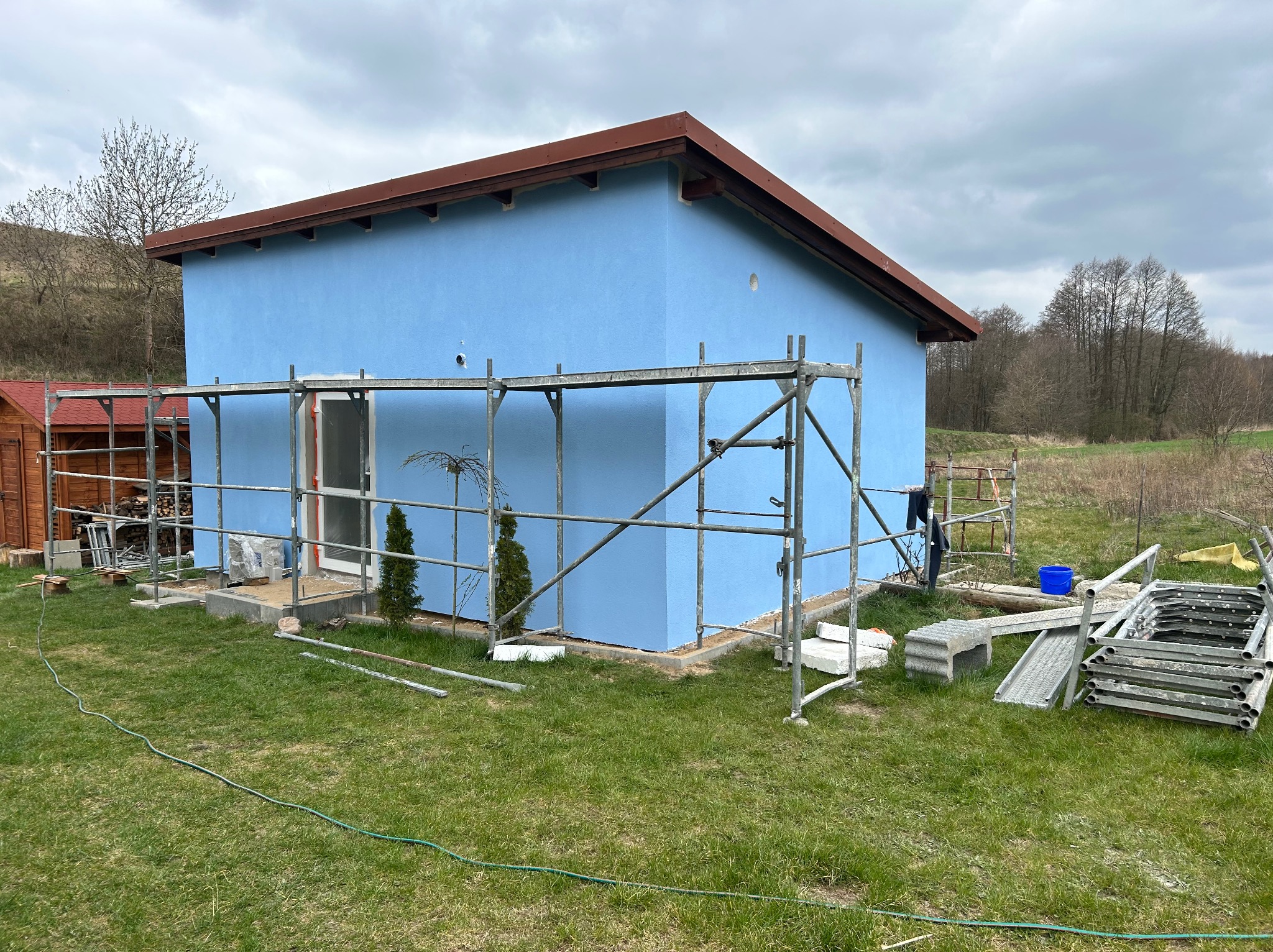 Niebieski dom z brązowym dachem w trakcie tynkowania, otoczony rusztowaniami i zieloną trawą. Widoczne narzędzia i materiały budowlane. Pochmurne niebo w tle.