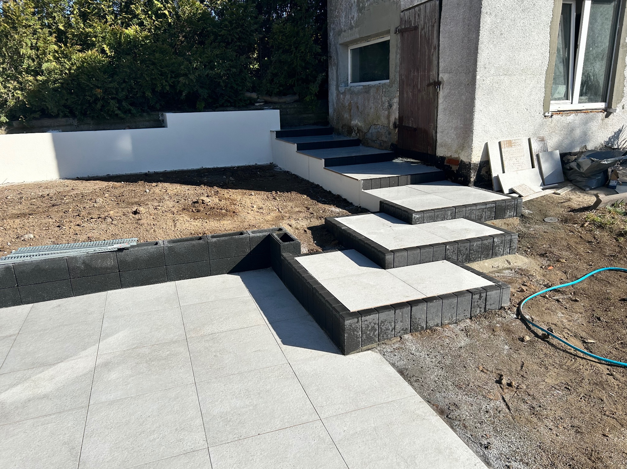 Ułożona jasnoszara kostka brukowa z ciemnymi obrzeżami tworząca ścieżkę i schody przy budynku. Widoczny trawnik, krzewy i fragment elewacji.