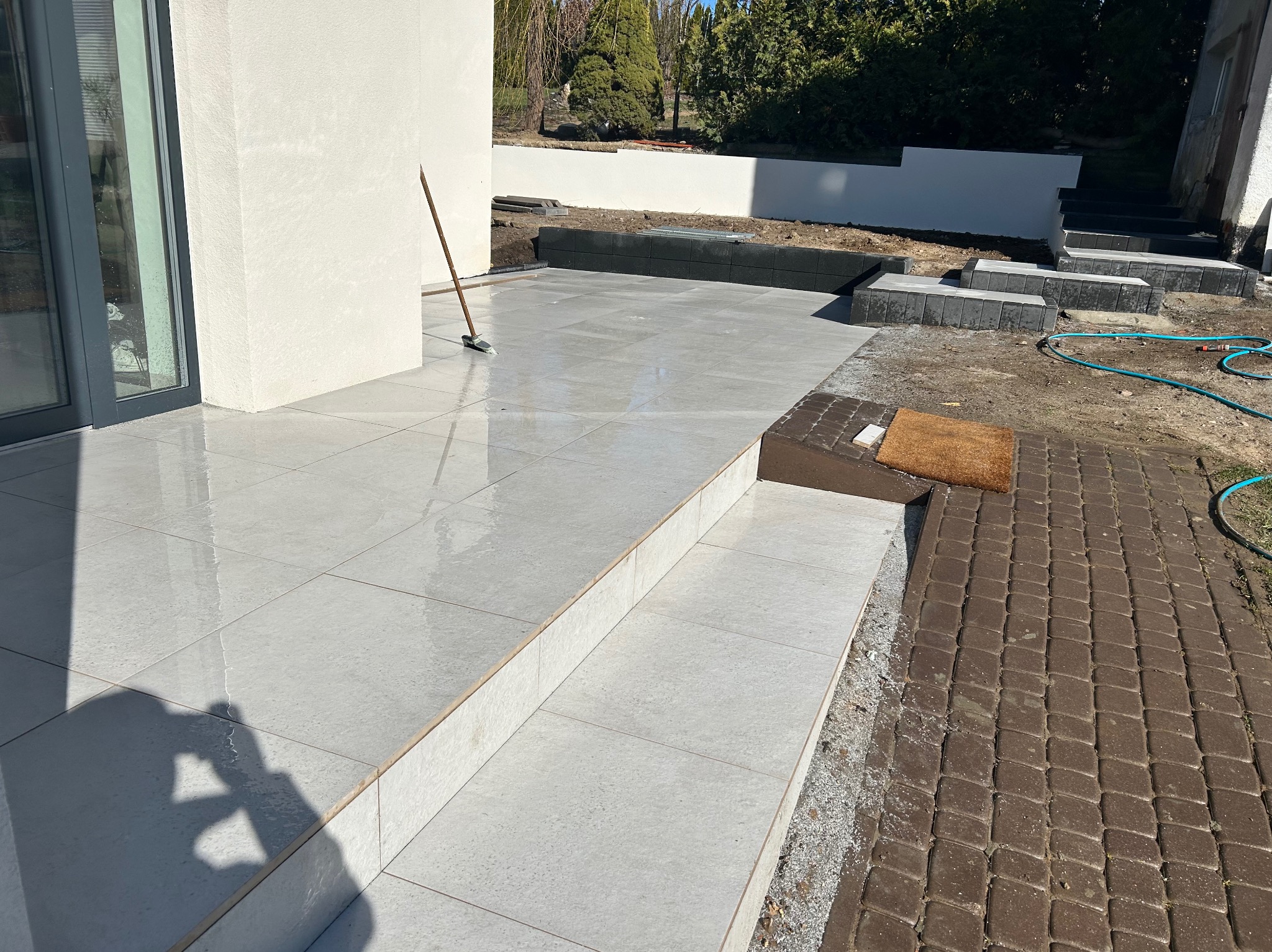 Jasnoszara, błyszcząca płytka na tarasie z dwoma stopniami. W tle widać fragment ogrodu i kostkę brukową. Na płytkach leży miotła.