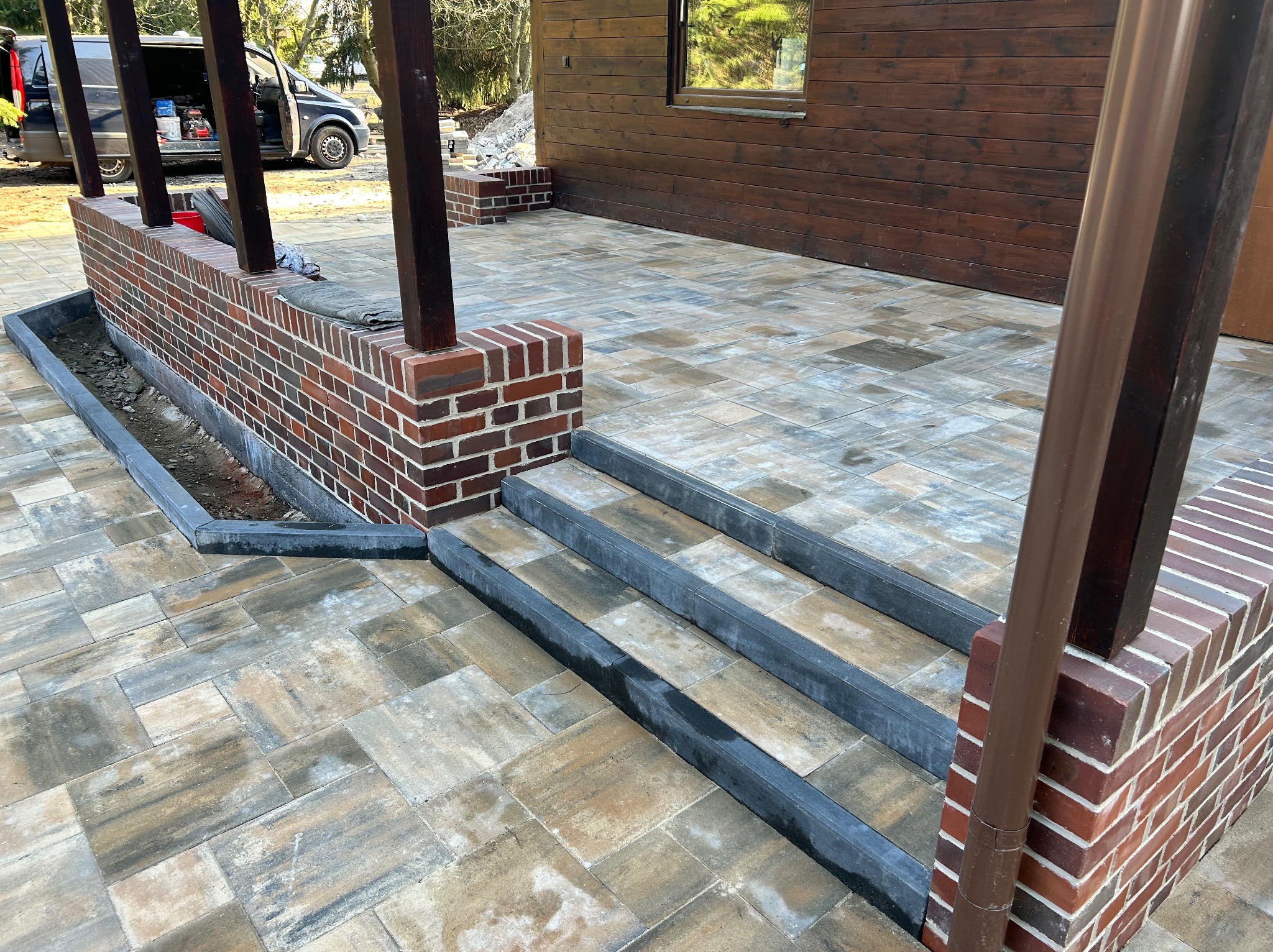 Ułożona kostka brukowa tworząca taras z ceglanymi elementami i schodami. Widoczna drewniana elewacja budynku w tle.