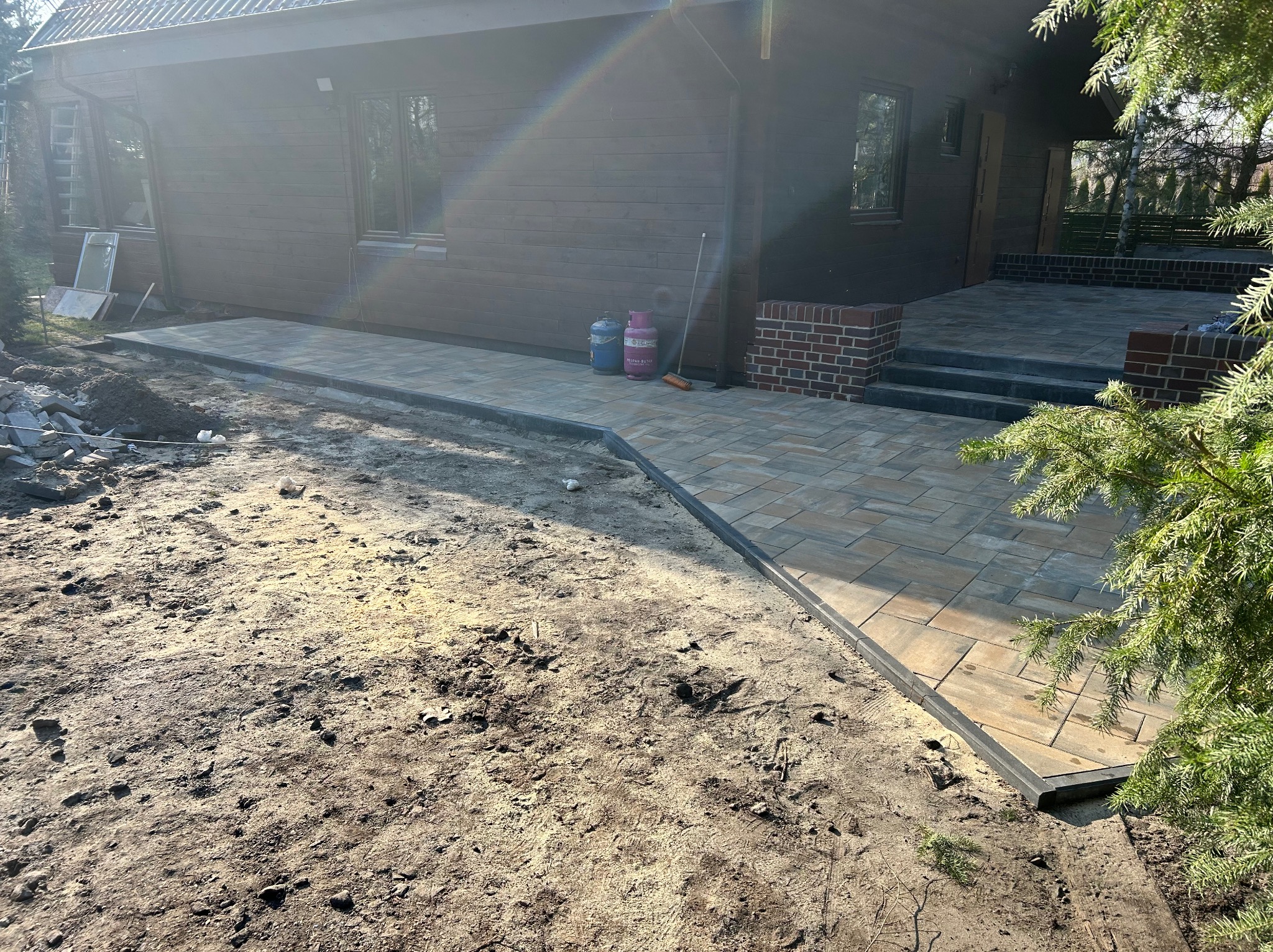 Ułożona kostka brukowa przed domem z drewnianą elewacją. Widoczne butle gazowe i częściowo ułożona nawierzchnia. Świecące słońce.