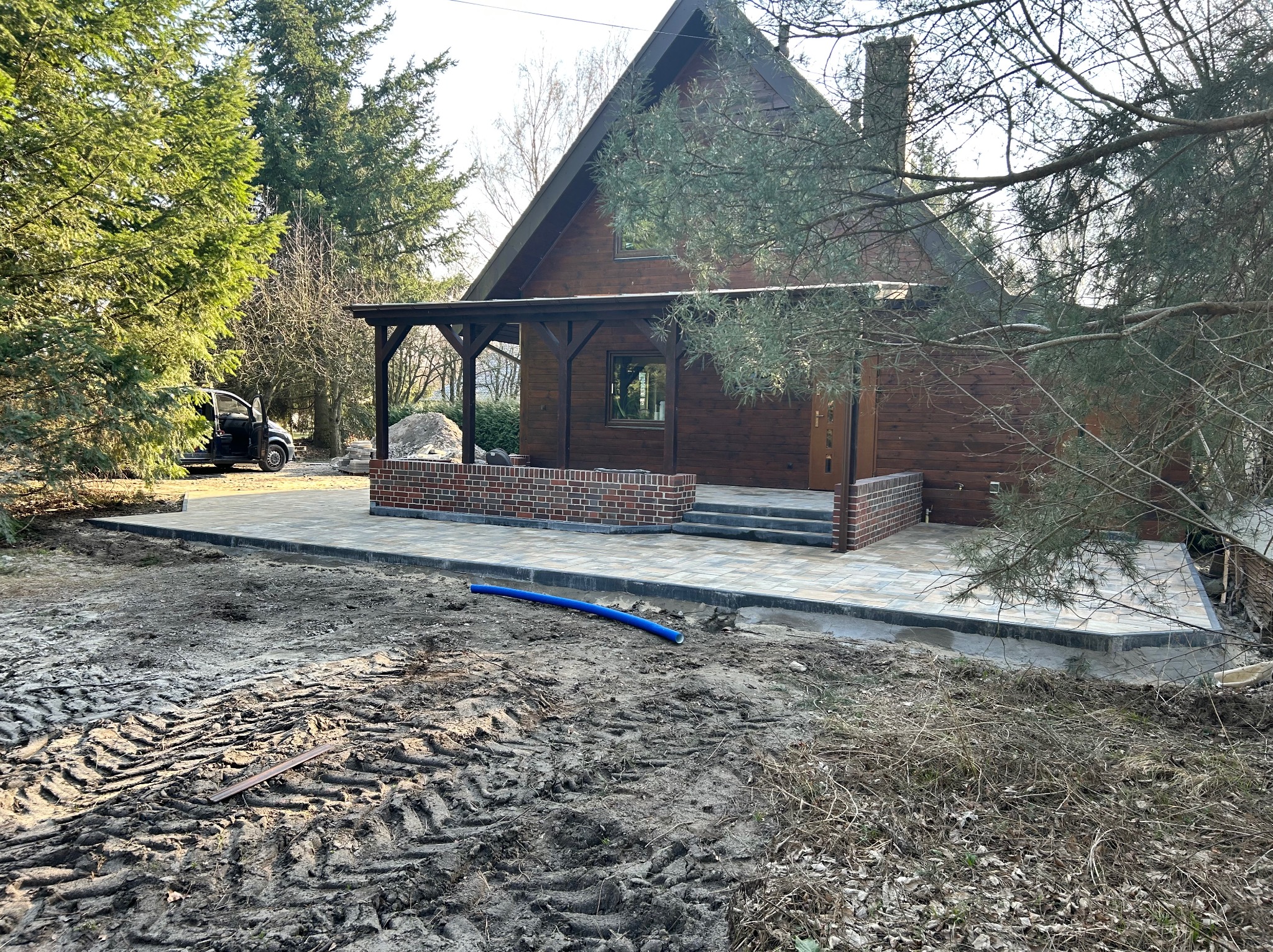 Drewniany dom z nową kostką brukową i ceglanym podmurówką tarasu, otoczony zielenią. Widoczne ślady opon na ziemi. Ciemny samochód w tle.