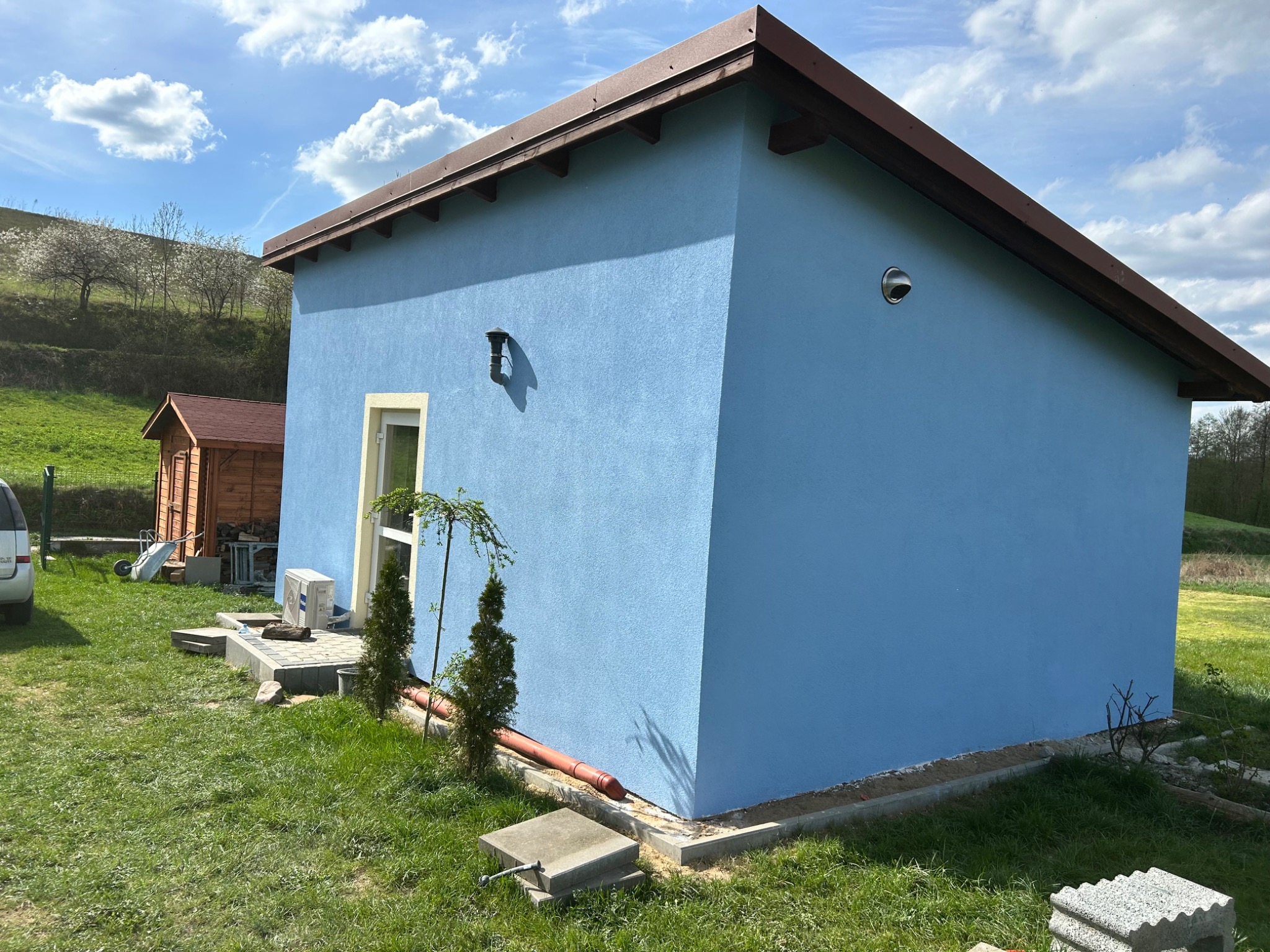 Niebieski budynek z brązowym dachem w słoneczny dzień, widoczne elementy instalacji na elewacji, trawnik i fragment drugiego budynku w tle.