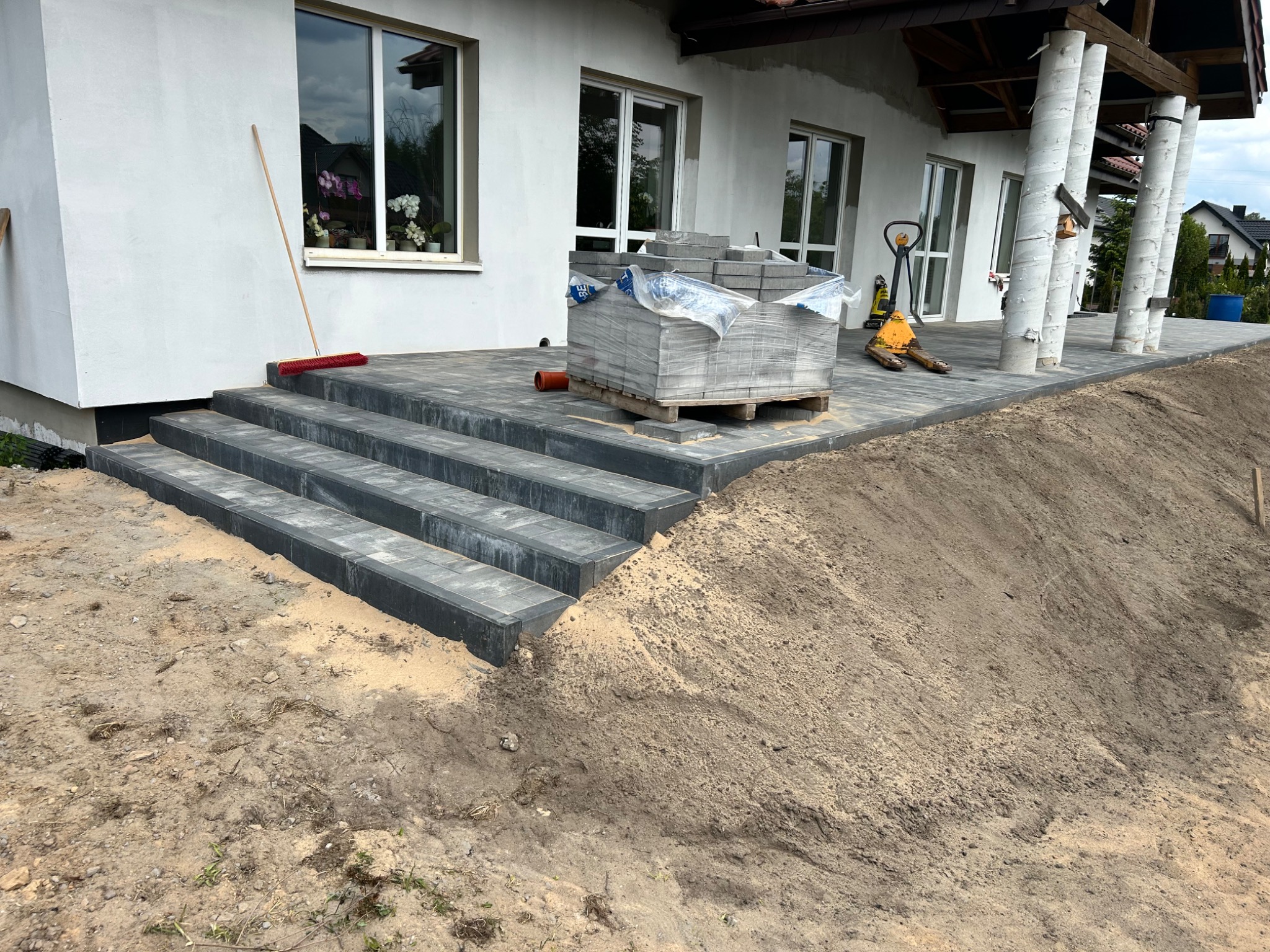 Ułożona kostka brukowa tworząca schody i taras przy domu. Paleta kostki, wózek paletowy, zamiatarka. Widok na białą elewację i kolumny podtrzymujące dach.