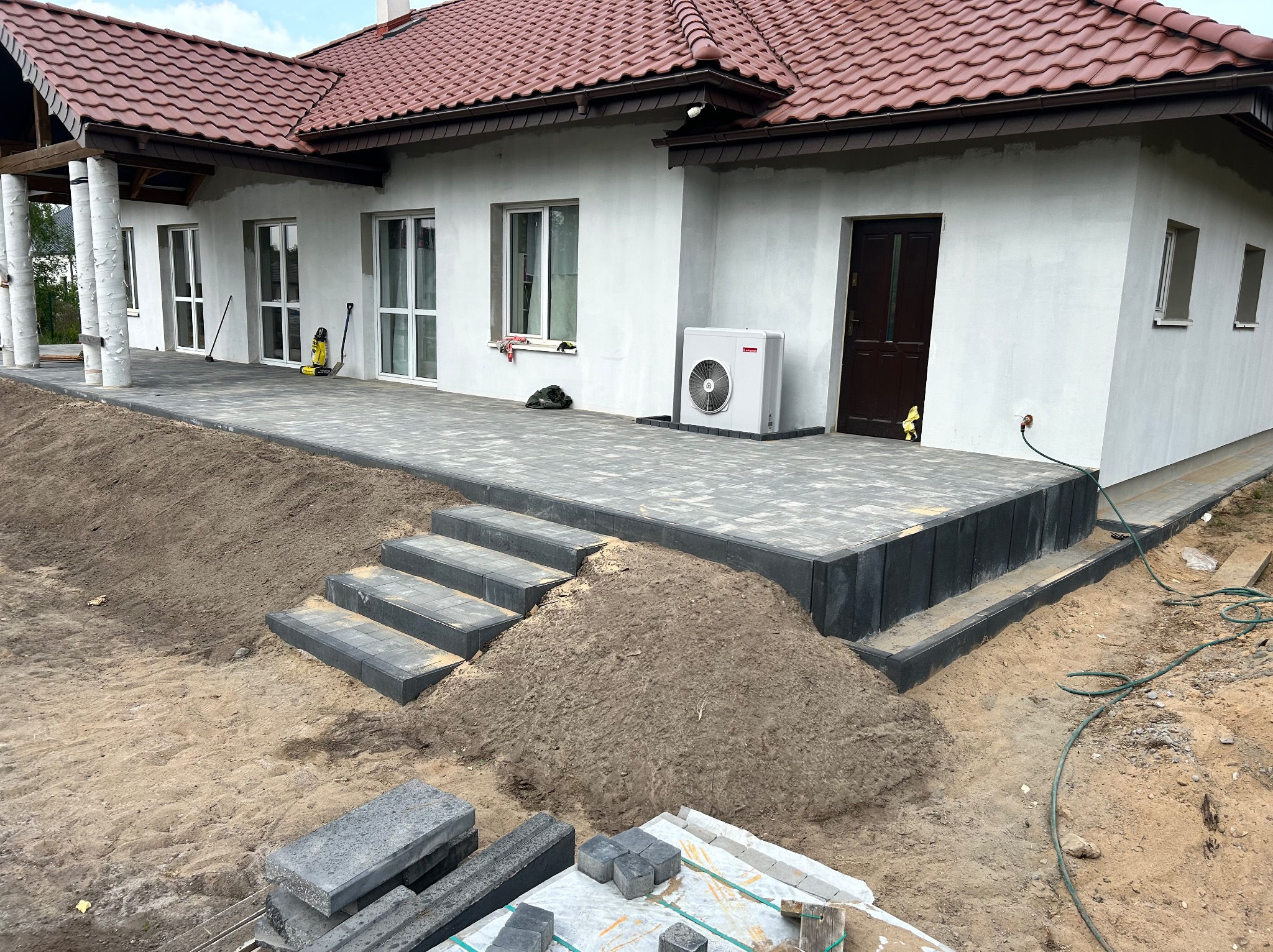 Ułożona kostka brukowa przed domem z brązowym dachem i białymi ścianami. Widoczne schody z kostki, trawnik i elementy wykończeniowe.