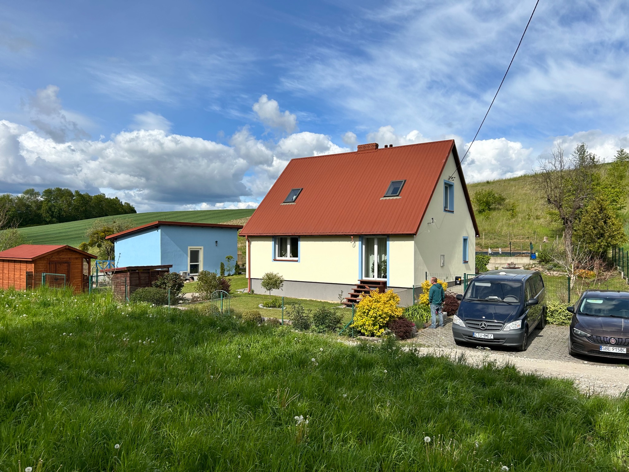 Jasny dom z czerwoną dachówką i oknami dachowymi, otoczony zielenią. Na podjeździe zaparkowane dwa samochody, w tle błękitny budynek i pagórkowaty krajobraz.