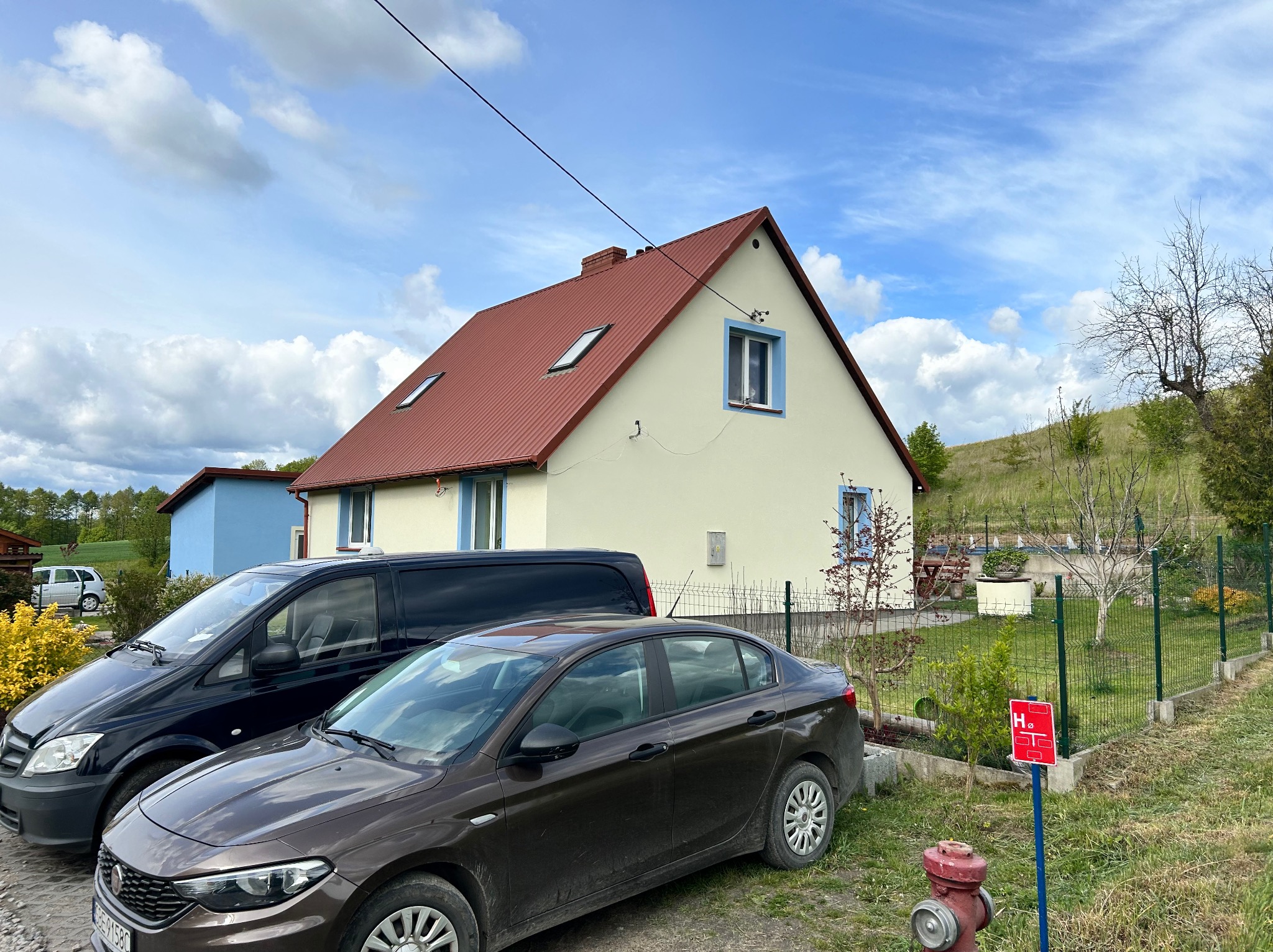 Jasny dom jednorodzinny z czerwoną dachówką i niebieskimi obramowaniami okien, otoczony zielenią i dwoma samochodami na pierwszym planie.