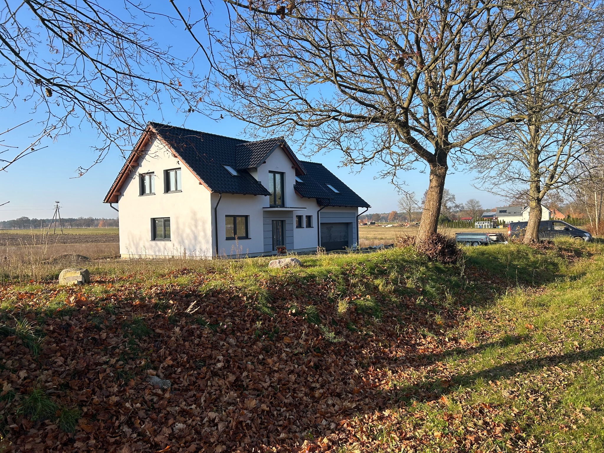 Nowoczesny dom jednorodzinny z garażem, wykończony w bieli, z ciemną dachówką, na tle jesiennego krajobrazu. Widoczne drzewa bez liści i opadłe liście.