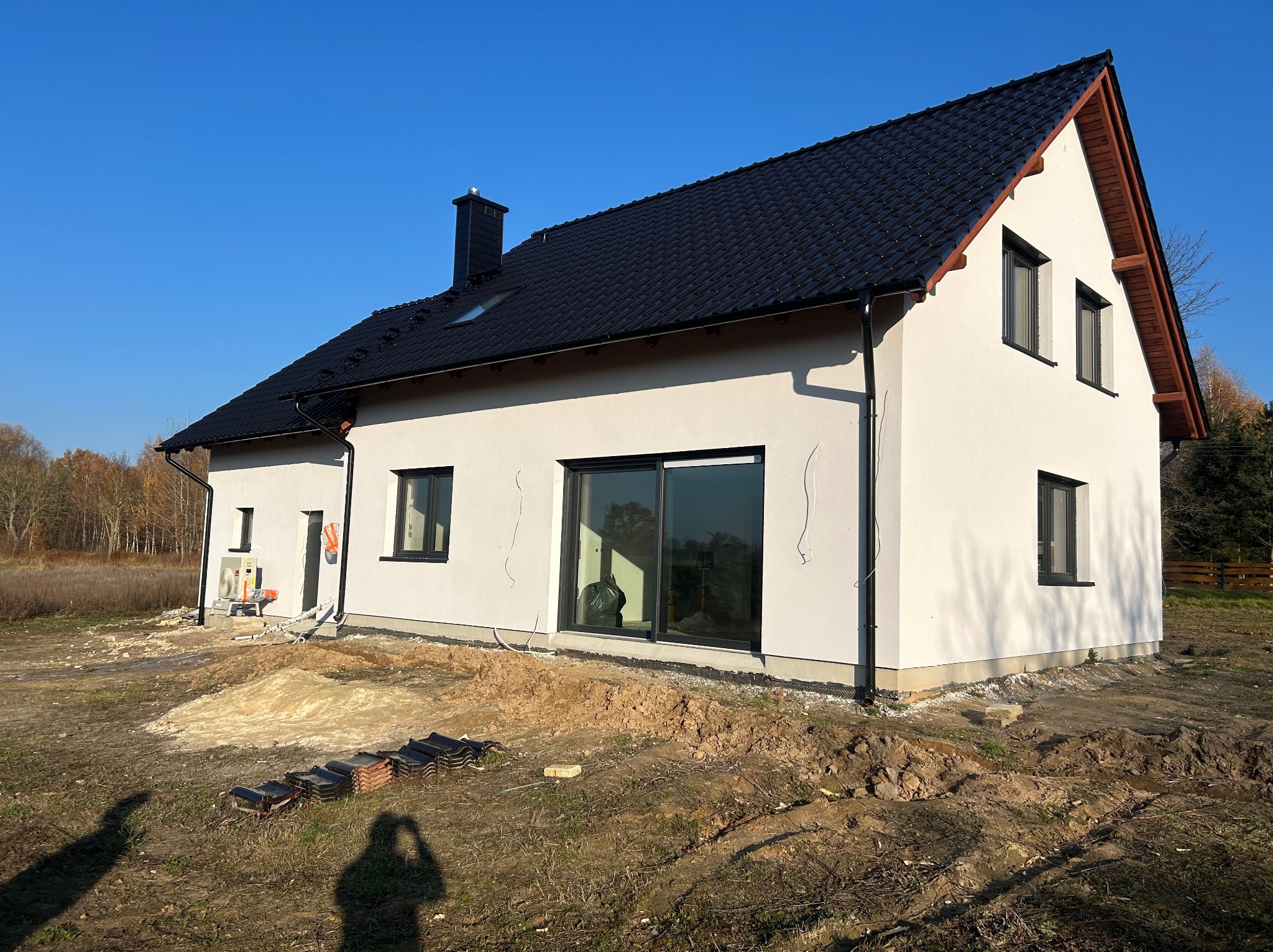 Nowoczesny dom jednorodzinny z białymi ścianami i czarnym dachem, w trakcie wykończenia terenu wokół budynku. Widoczne ułożone dachówki i klimatyzator na elewacji.