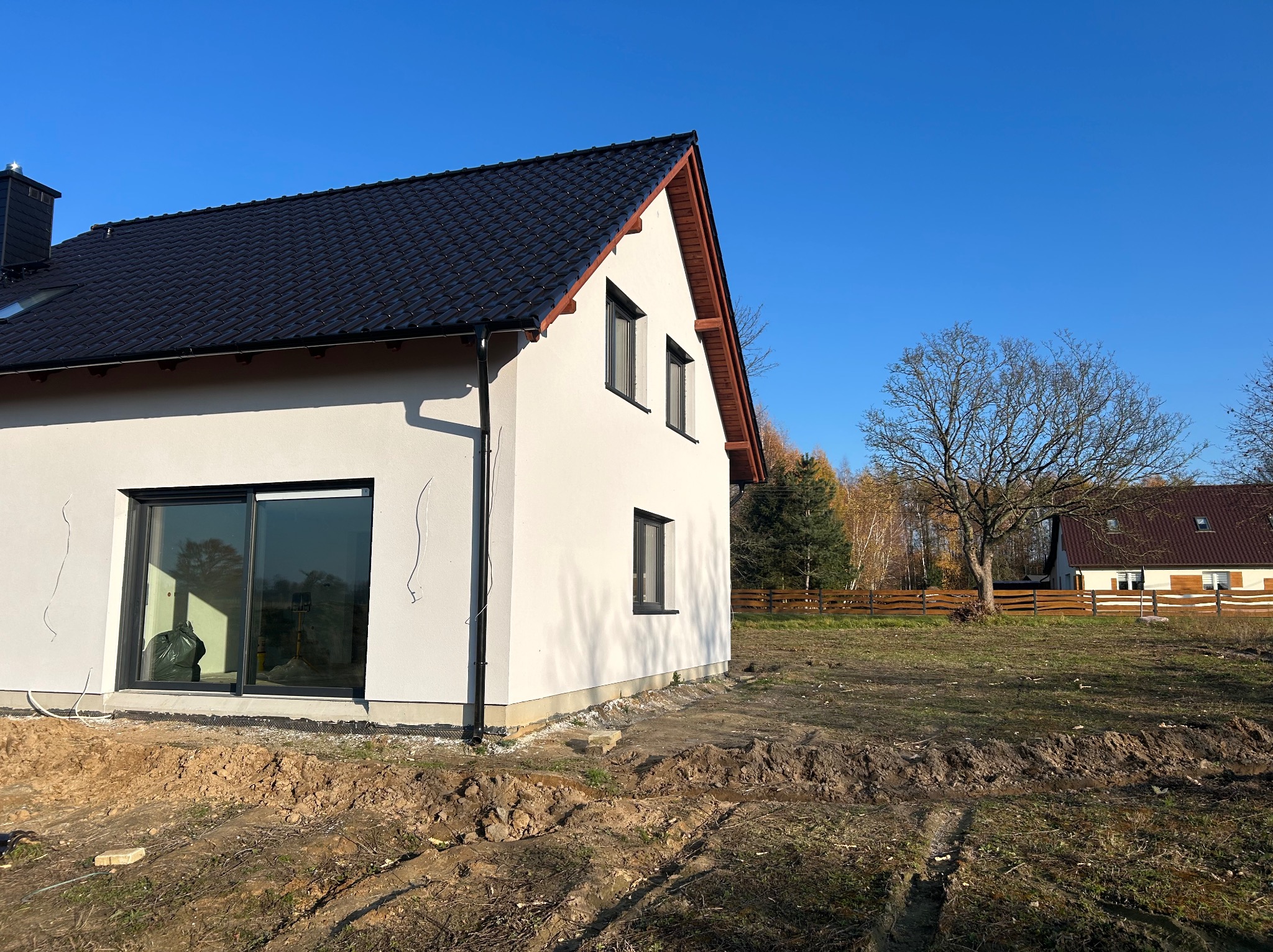 Nowoczesny dom jednorodzinny z czarnym dachem i białymi ścianami, duże okno przesuwne, widok z zewnątrz na niezagospodarowany teren.