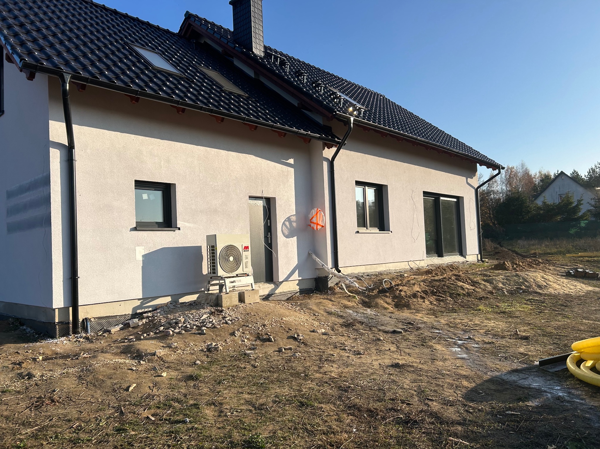 Nowoczesny dom z czarną dachówką i klimatyzatorem na zewnątrz. Widoczne prace ziemne wokół budynku i rury. Jasne tynki elewacyjne i okna w ciemnych ramach.