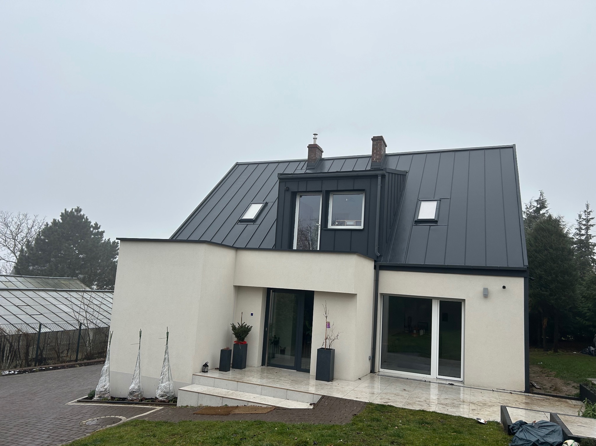 Nowoczesny dom z ciemnym dachem i jasną elewacją w pochmurny dzień. Widoczne okna dachowe, kominy i rośliny w donicach przy wejściu.