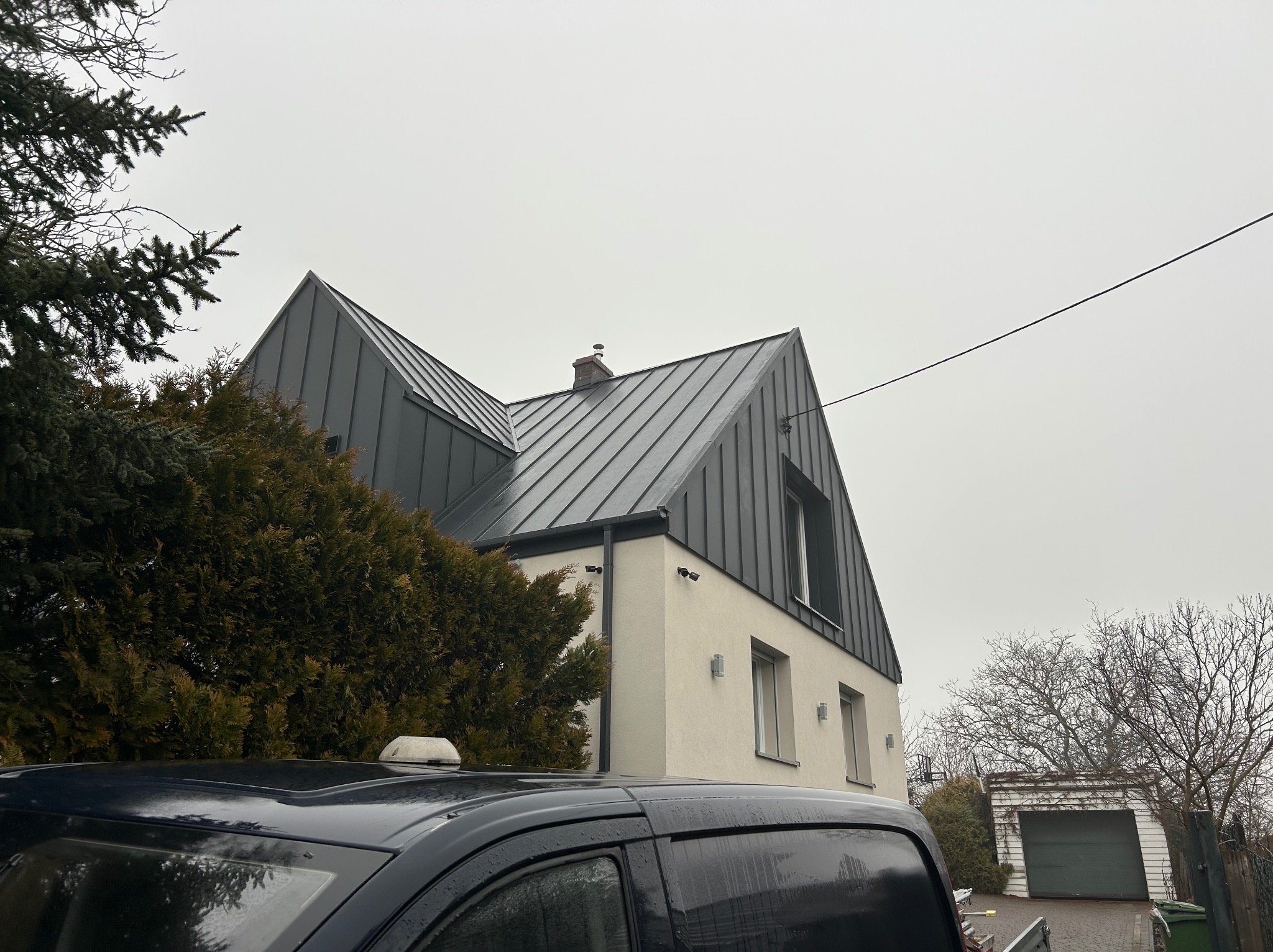 Nowoczesny dom z szarą blachą trapezową na dachu, kremową elewacją i minimalistycznymi lampami zewnętrznymi, częściowo zasłonięty przez drzewa i samochód.