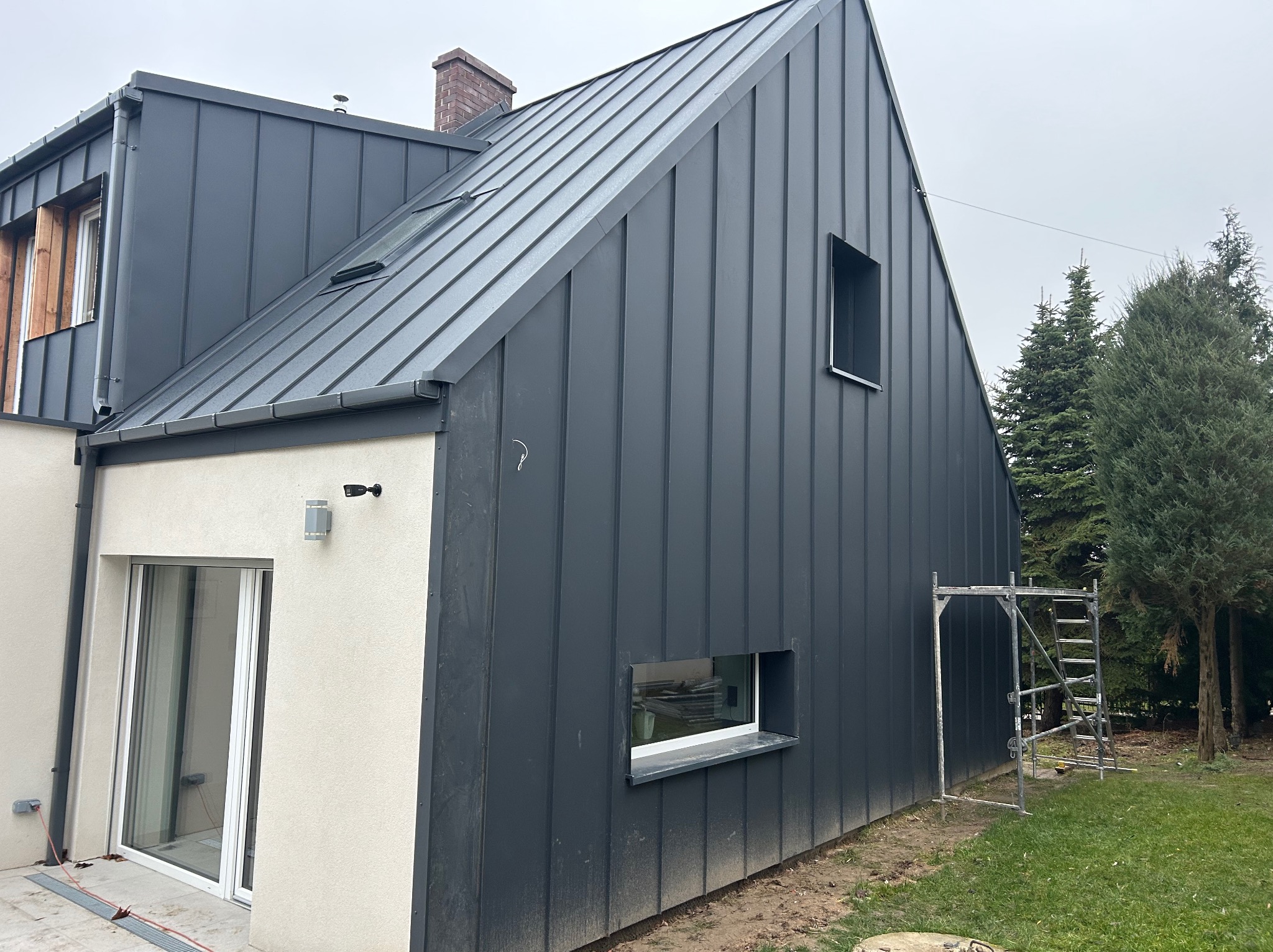 Nowoczesny dom z ciemną, metalową elewacją i jasnymi elementami wykończeniowymi. Widoczne rusztowanie, sugerujące prace wykończeniowe. Ujęcie z boku budynku.