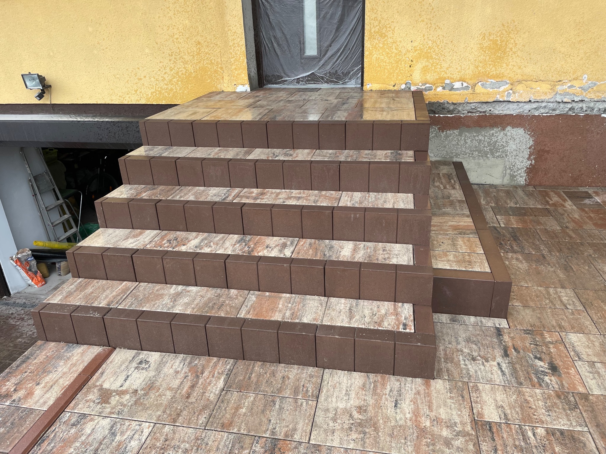 Ułożone schody wejściowe z kostki brukowej w odcieniach brązu i beżu, prowadzące do domu z żółtą elewacją. Widoczne detale wykończenia i wzór kostki.