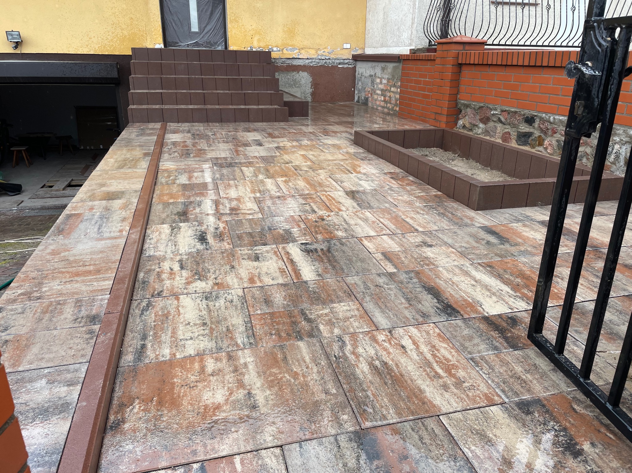 Ułożona kostka brukowa w odcieniach brązu i beżu, tworząca ścieżkę i schody przed budynkiem. Widoczny fragment muru z cegły i metalowe ogrodzenie.
