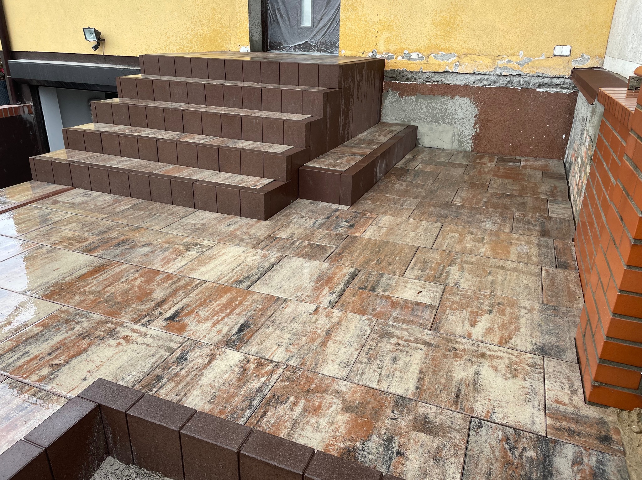 Ułożona kostka brukowa w odcieniach brązu i beżu, tworząca schody i chodnik przed budynkiem. Widoczne detale wykończenia i świeżo położona nawierzchnia.