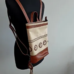 Beżowo-brązowy skórzany plecak z etnicznymi zdobieniami na czarnym manekinie, wiszący brelok z frędzlami.