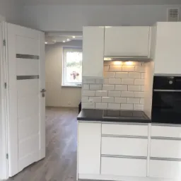 Nowoczesna, jasna kuchnia z białymi szafkami i czarnym blatem, widoczna przez otwarte drzwi z trzema poziomymi, srebrnymi paskami, z wnętrzem mieszkania w tle.