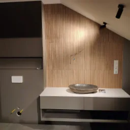 Nowoczesna łazienka w skosach: szafka pod umywalkę z blatem, kamienna misa umywalkowa, obudowa WC w zabudowie, drewniana okładzina ścienna z pionowymi listwami, widoczne przewody instalacji...