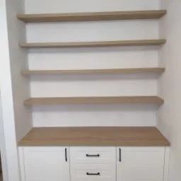 Biała zabudowa meblowa z trzema szufladami i dwojgiem drzwiczek, zwieńczona blatem w kolorze jasnego drewna, nad którą znajdują się cztery drewniane półki na białej ścianie.